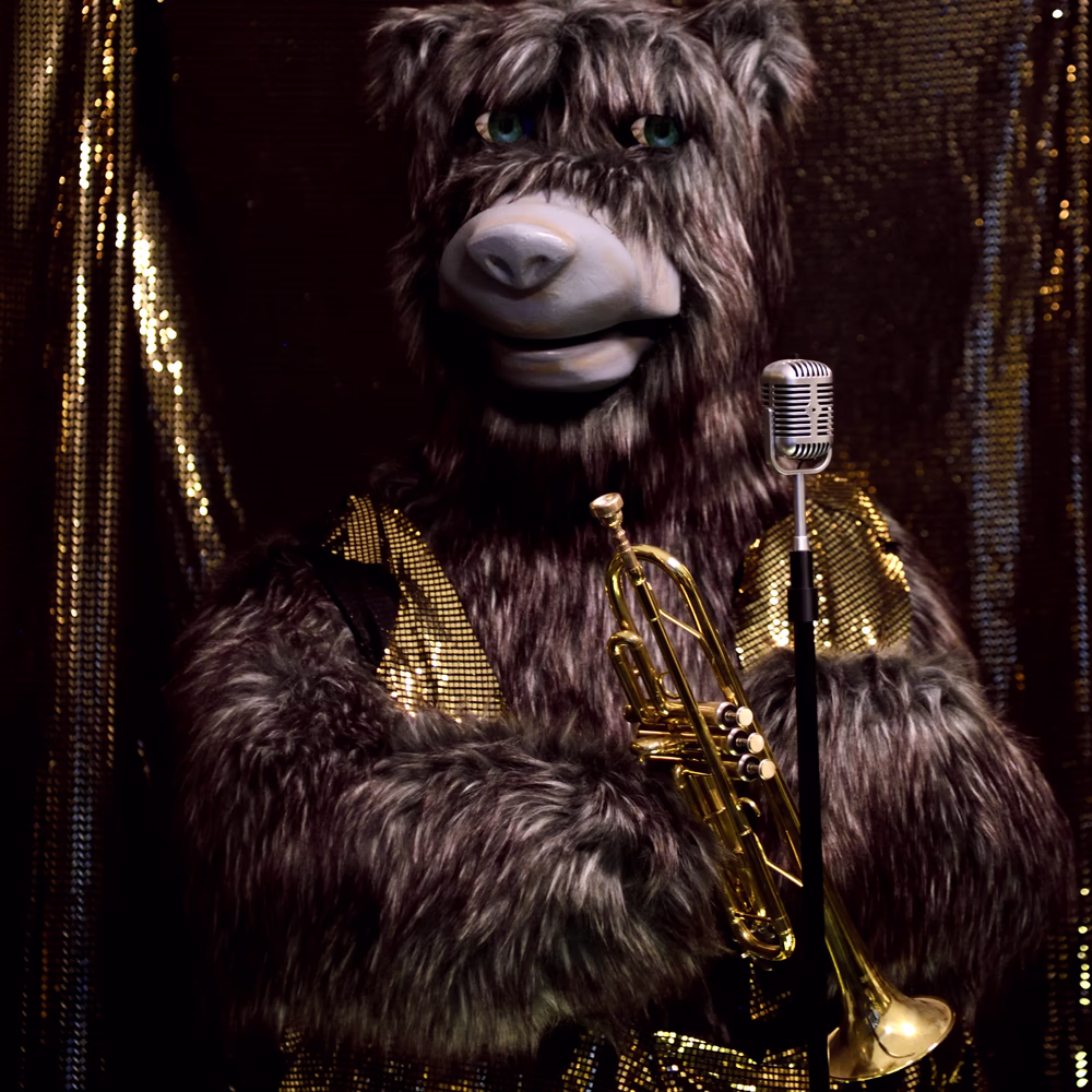 Orso animatronico grigio-marrone della Bear Band con occhi verdi, vestito dorato luccicante, tiene tromba dorata con microfono vintage davanti. Sfondo con tende metalliche dorate