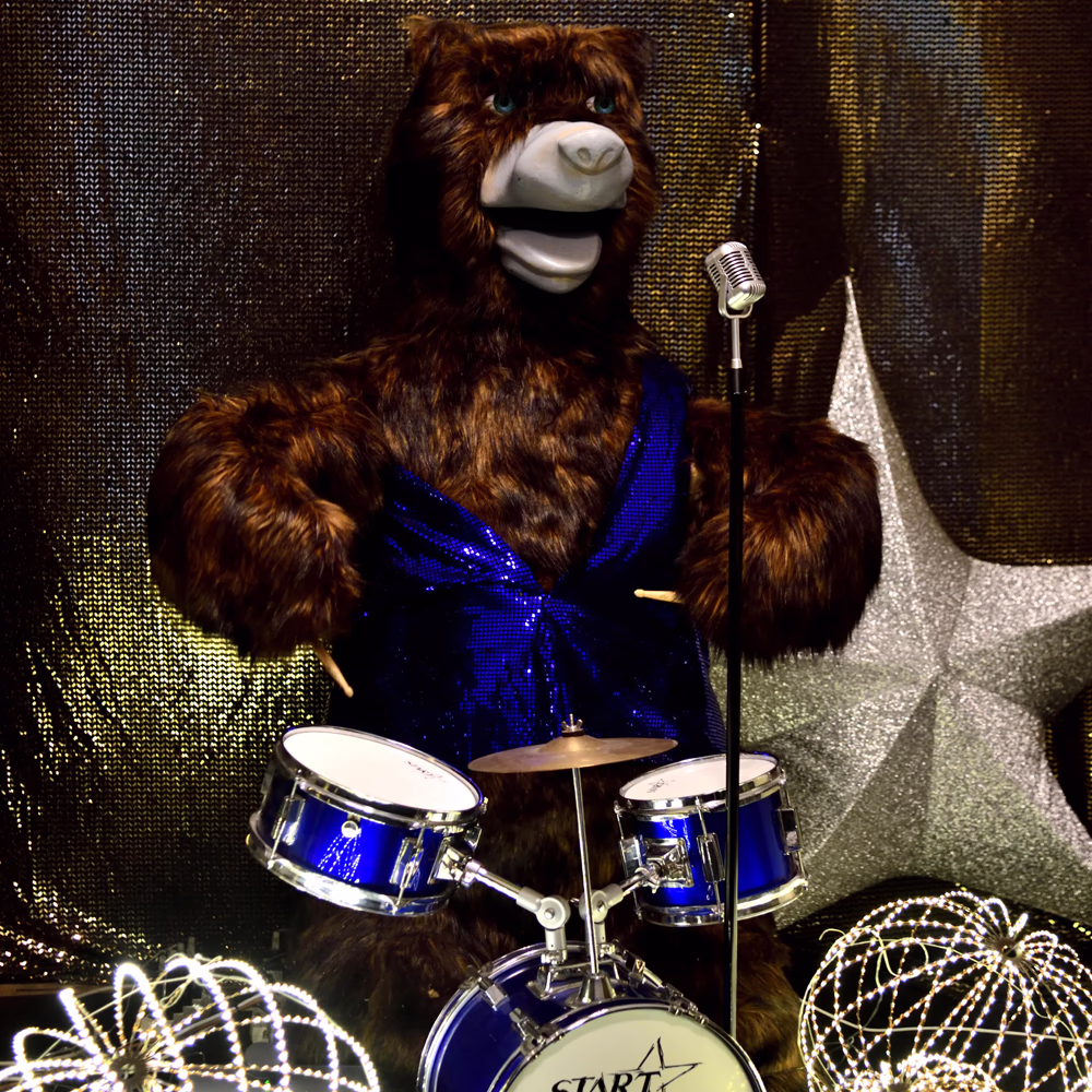 Orso animatronico marrone della Bear Band con pelliccia folta, vestito blu luccicante, seduto dietro batteria blu con microfono vintage. Sfondo con tende metalliche dorate e stelle decorative