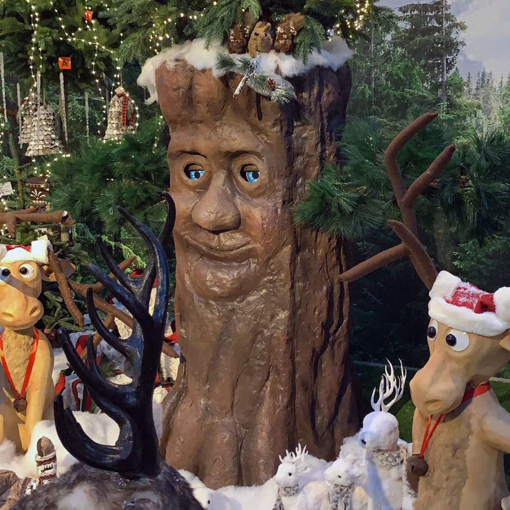 Natalbero, albero animato Menart con volto scolpito e occhi LED azzurri, circondato da renne scenografiche in villaggio natalizio innevato.