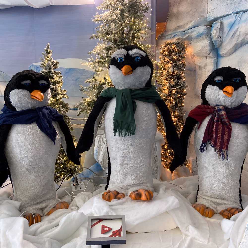 Trio Pinguini scenografici con occhi LED in paesaggio artico Menart, ambientazione con neve artificiale e alberi illuminati per villaggio natalizio completo.