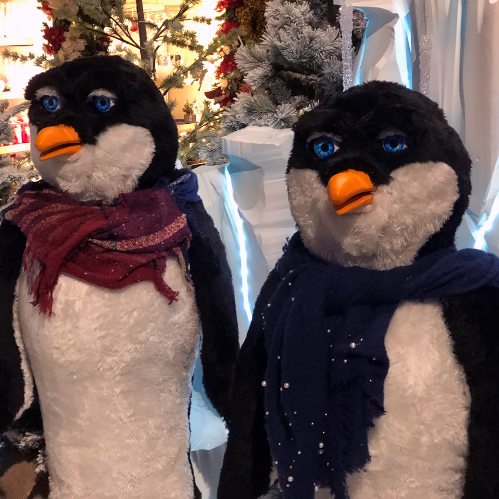 Pinguini Menart con occhi LED blu e sciarpe colorate, installazione scenografica natalizia innevata con illuminazione atmosferica per esperienza invernale immersiva.