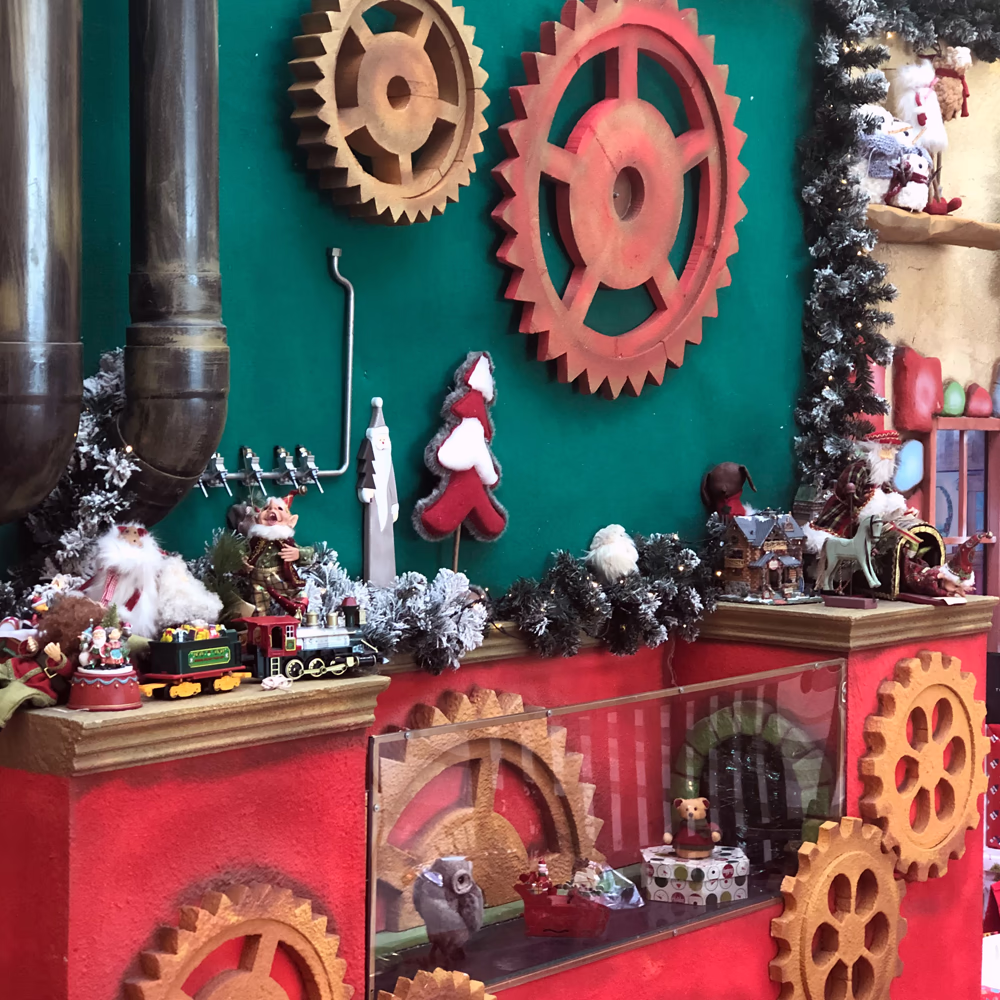 Scenografia meccanica con sistema di ingranaggi funzionali, elementi industriali vintage, illuminazione LED decorativa e esposizione di giocattoli tradizionali in ambientazione natalizia suggestiva.