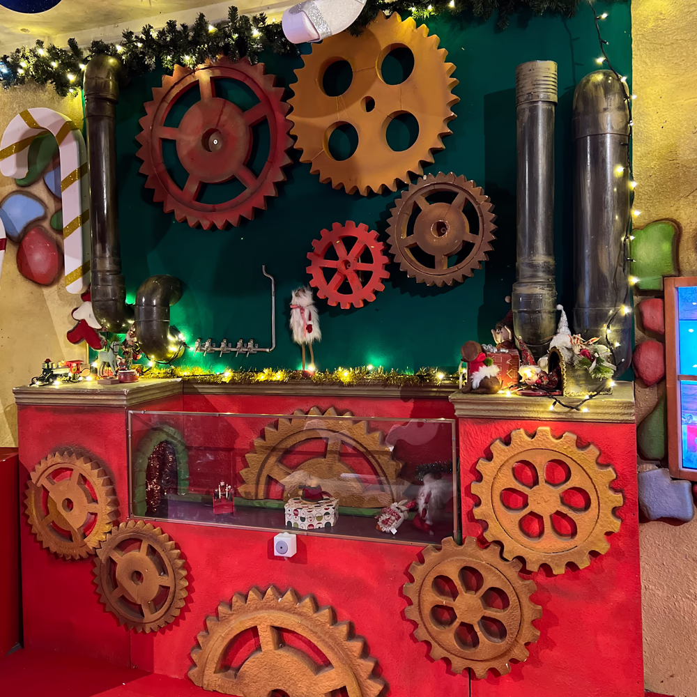 Allestimento scenografico industriale con ingranaggi meccanici giganti, tubi metallici e vetrina espositiva rossa, creando un'atmosfera di meccanica vintage per "La Fabbrica Dei Giocattoli".