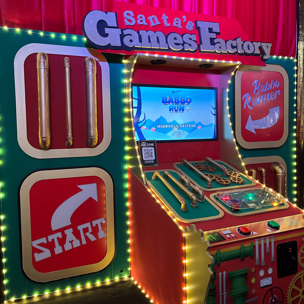 Installazione gaming interattiva "Santa's Games Factory" con console arcade natalizia, schermo digitale, controlli fisici e illuminazione decorativa per esperienza ludica immersiva e coinvolgente.