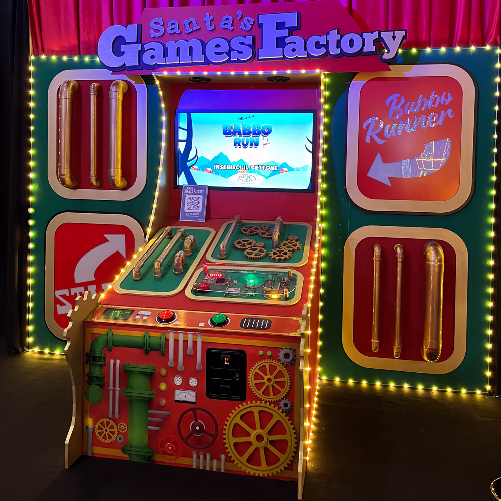 Installazione gaming interattiva "Santa's Games Factory" con console arcade tematizzata, schermi digitali, controlli meccanici industriali e illuminazione decorativa per esperienza ludica immersiva e coinvolgente natalizia.