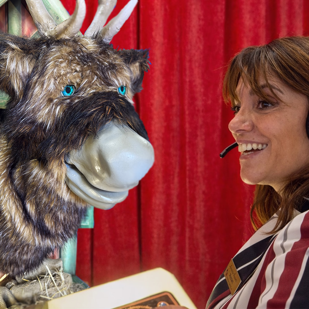 Smiling woman with headphone and badge talks with a figure with the head of a furry animal with horns and blue eyes.