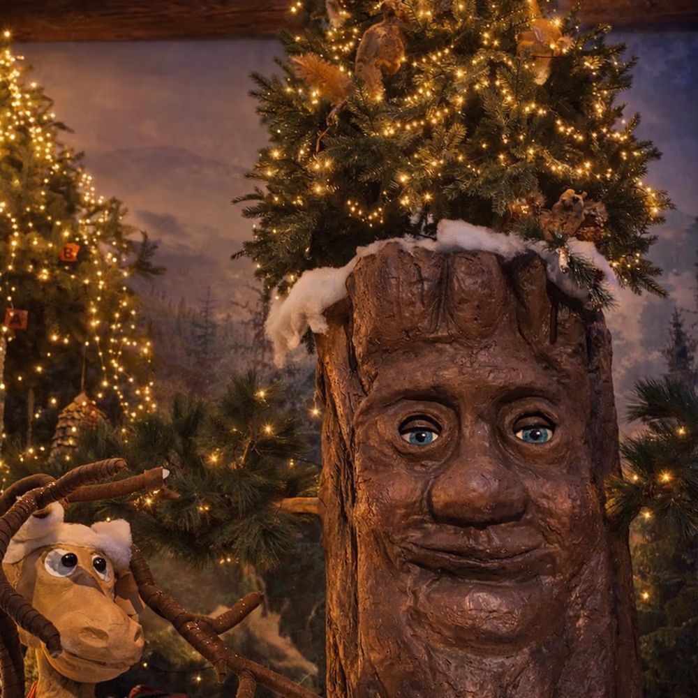 Christmas tree with smiling wooden face and bright decorations, with a reindeer figure in the lower left corner.