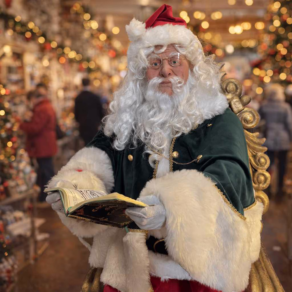 Babbo Natale con barba bianca in giacca verde e rossa tiene un libro aperto in una stanza decorata per Natale.