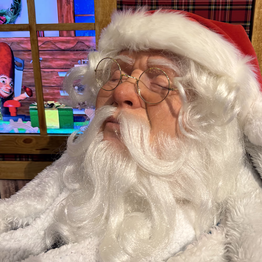 Babbo Natale con barba bianca, occhiali tondi e cappello rosso, seduto vicino a una finestra decorata.