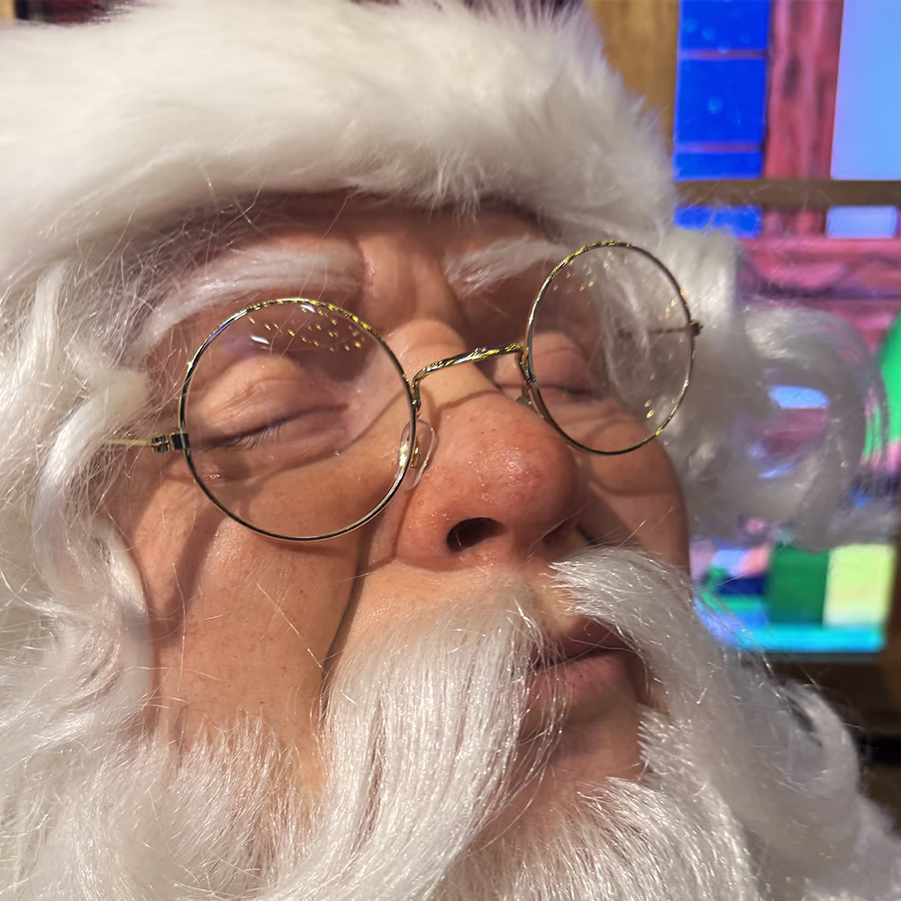 Primo piano del volto di Babbo Natale con capelli bianchi, barba folta e occhiali tondi dorati.