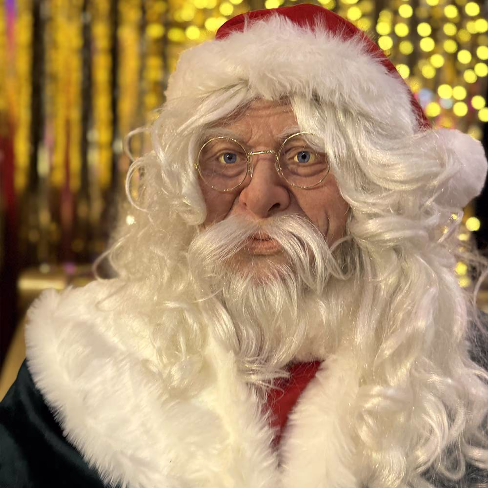 Primo piano di Babbo Natale con barba bianca, occhiali rotondi e sfondo sfocato di luci natalizie.