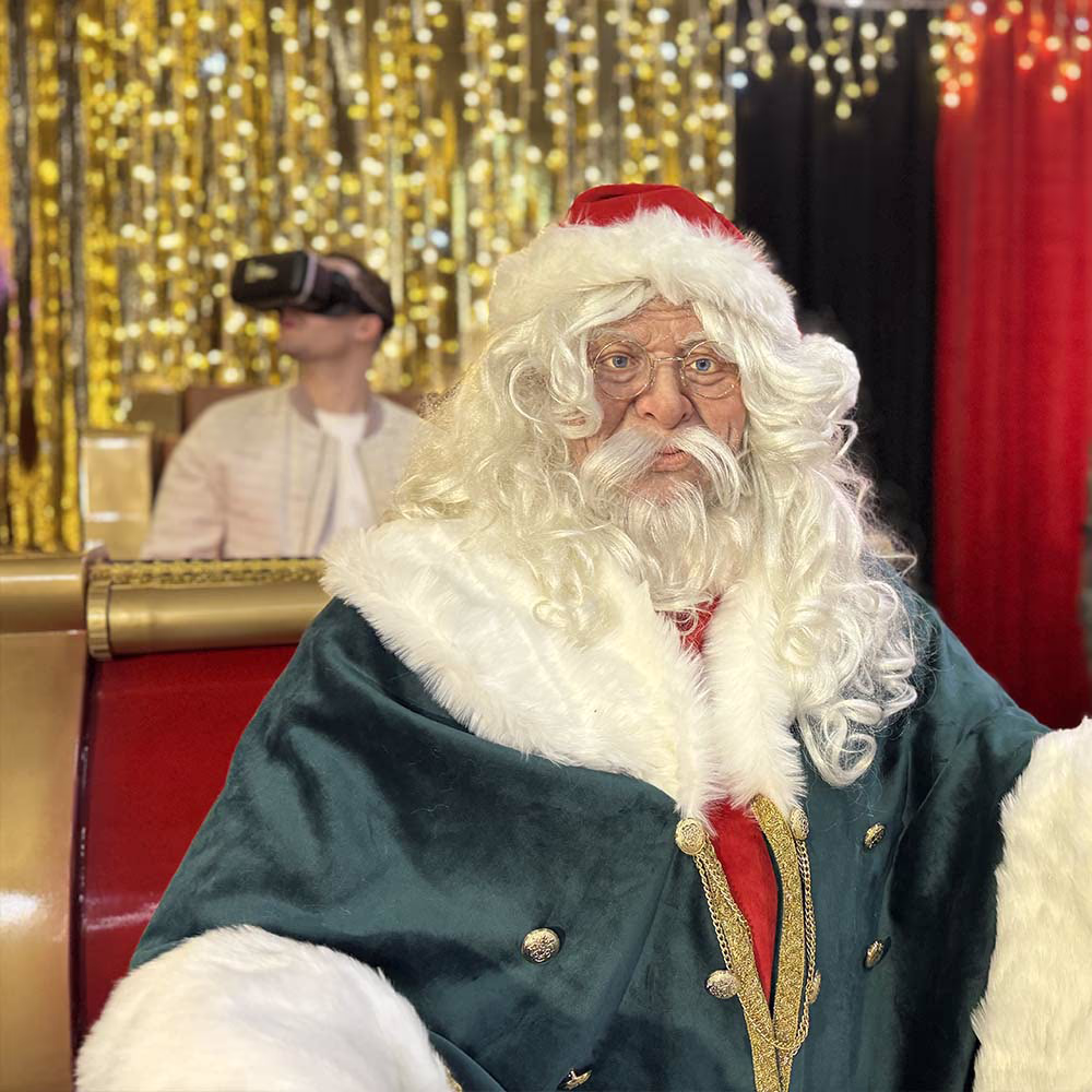 Babbo Natale con abito verde, barba bianca e occhiali davanti a una persona che usa visore VR.