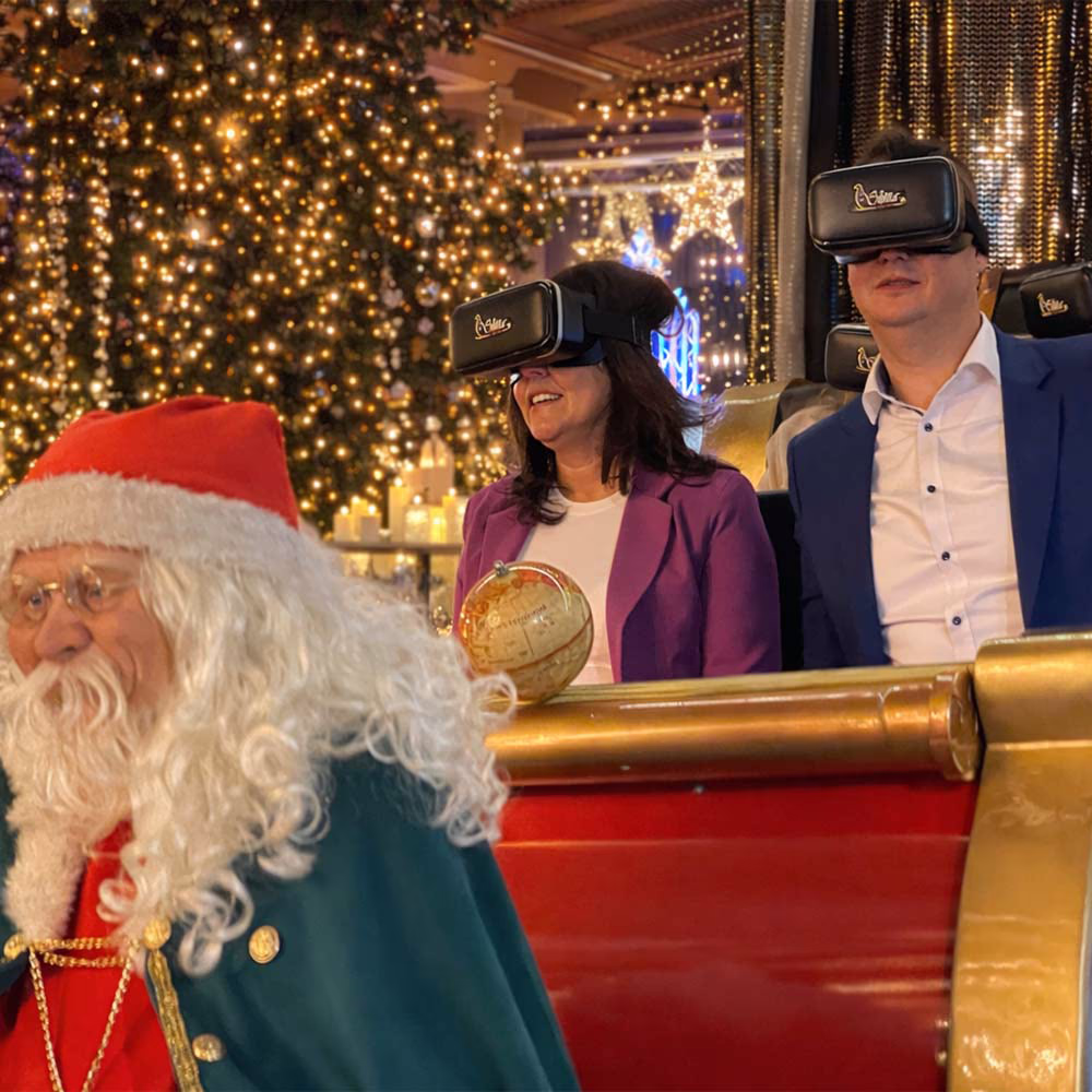 Due persone indossano visori VR sedute su una slitta festiva davanti a un grande albero di Natale illuminato.