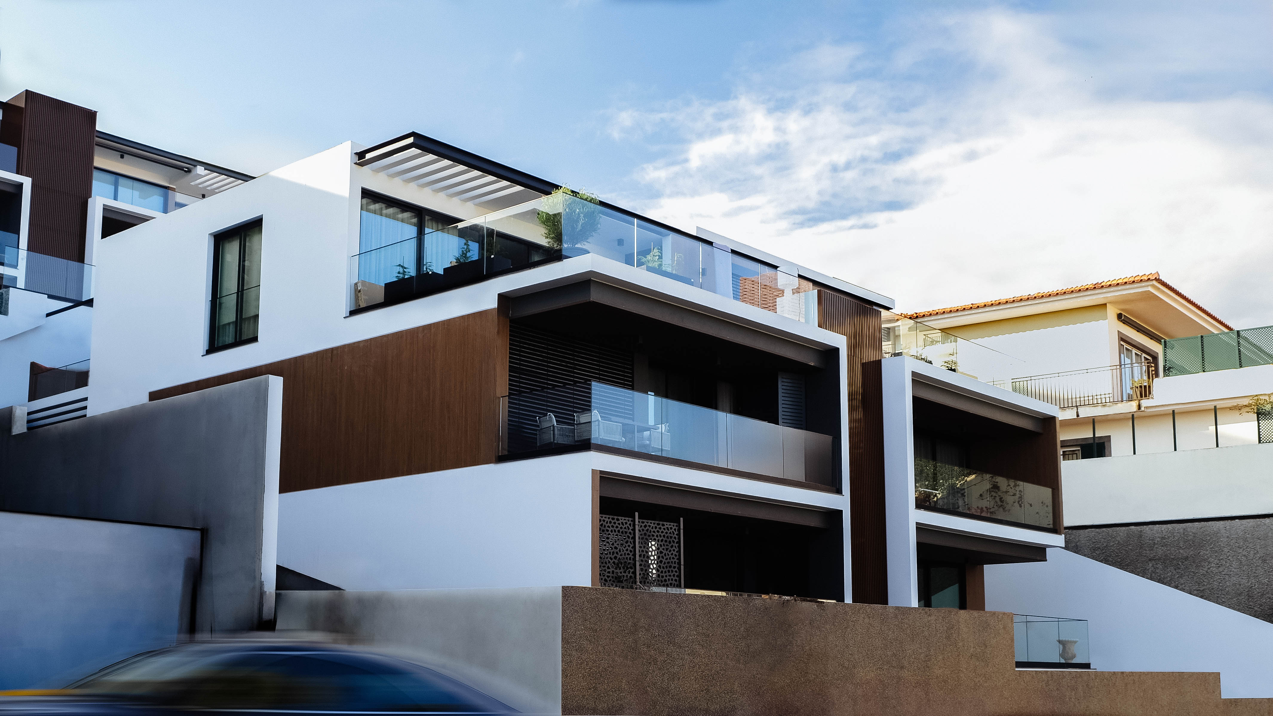 Uma moradia moderna de luxo na Madeira, construída em socalcos, com fachada branca e detalhes em madeira. A casa apresenta grandes janelas, varandas com parapeitos de vidro e uma garagem integrada. Um carro em movimento pode ser visto em primeiro plano.