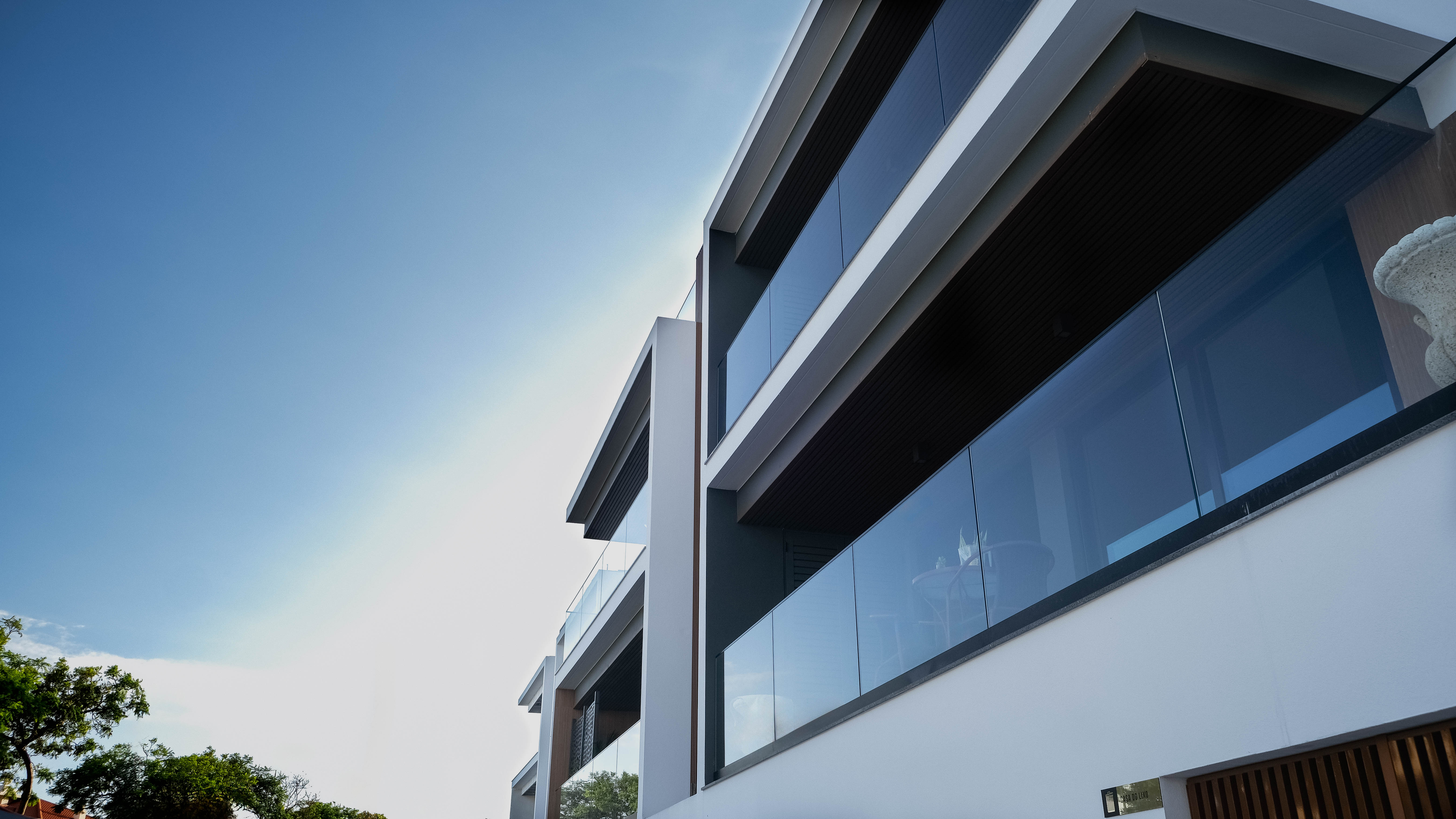 Vista de baixo para cima de um edifício de apartamentos moderno, com varandas com parapeitos de vidro e um céu azul claro. A arquitetura é minimalista, com linhas retas e detalhes em madeira.