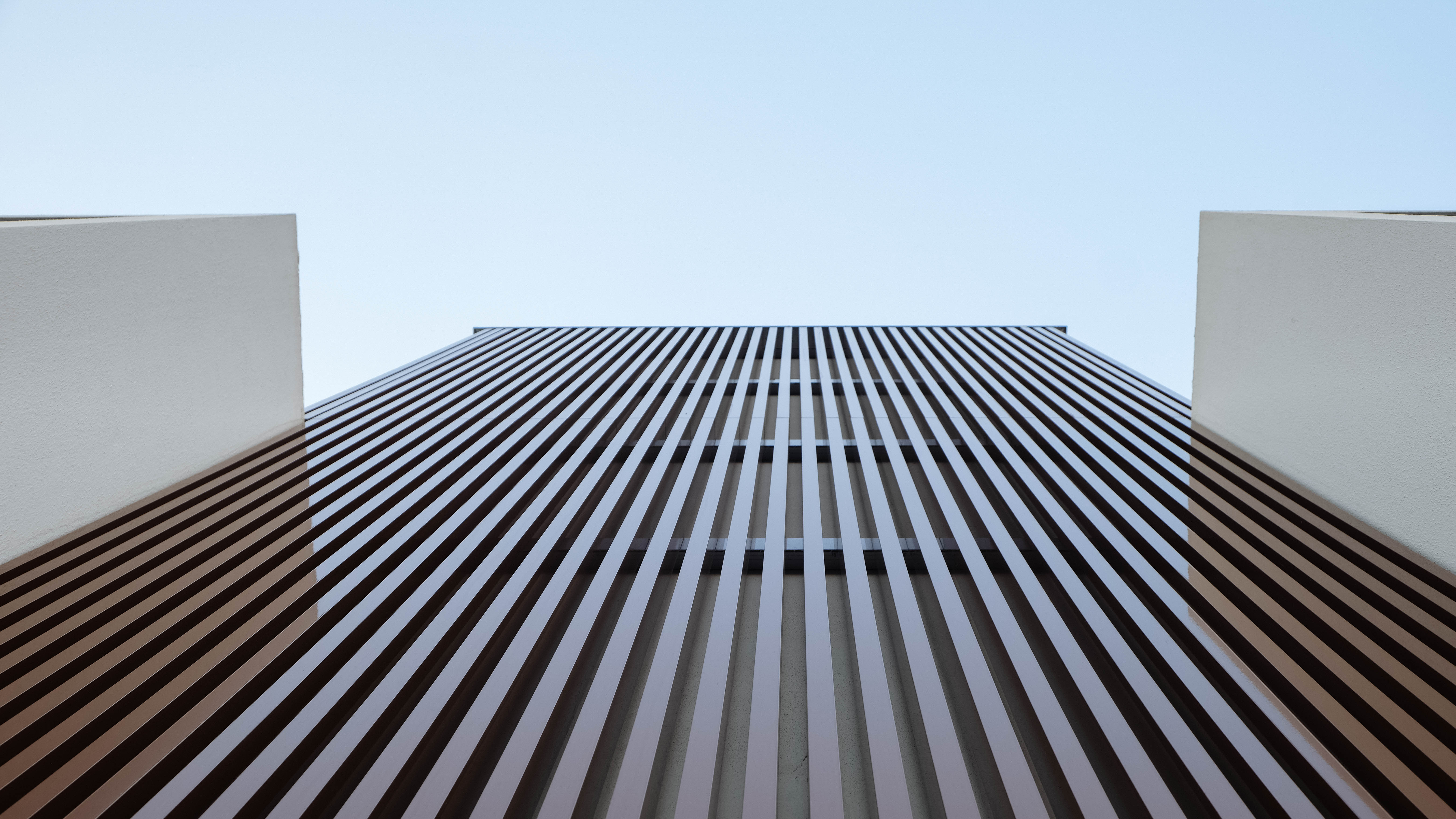 Detalhe da fachada de um edifício moderno, com uma parede coberta por painéis de ripas de madeira escura, criando um padrão vertical. A imagem é vista de baixo para cima, com as linhas a convergir para o topo, contra um céu azul claro.