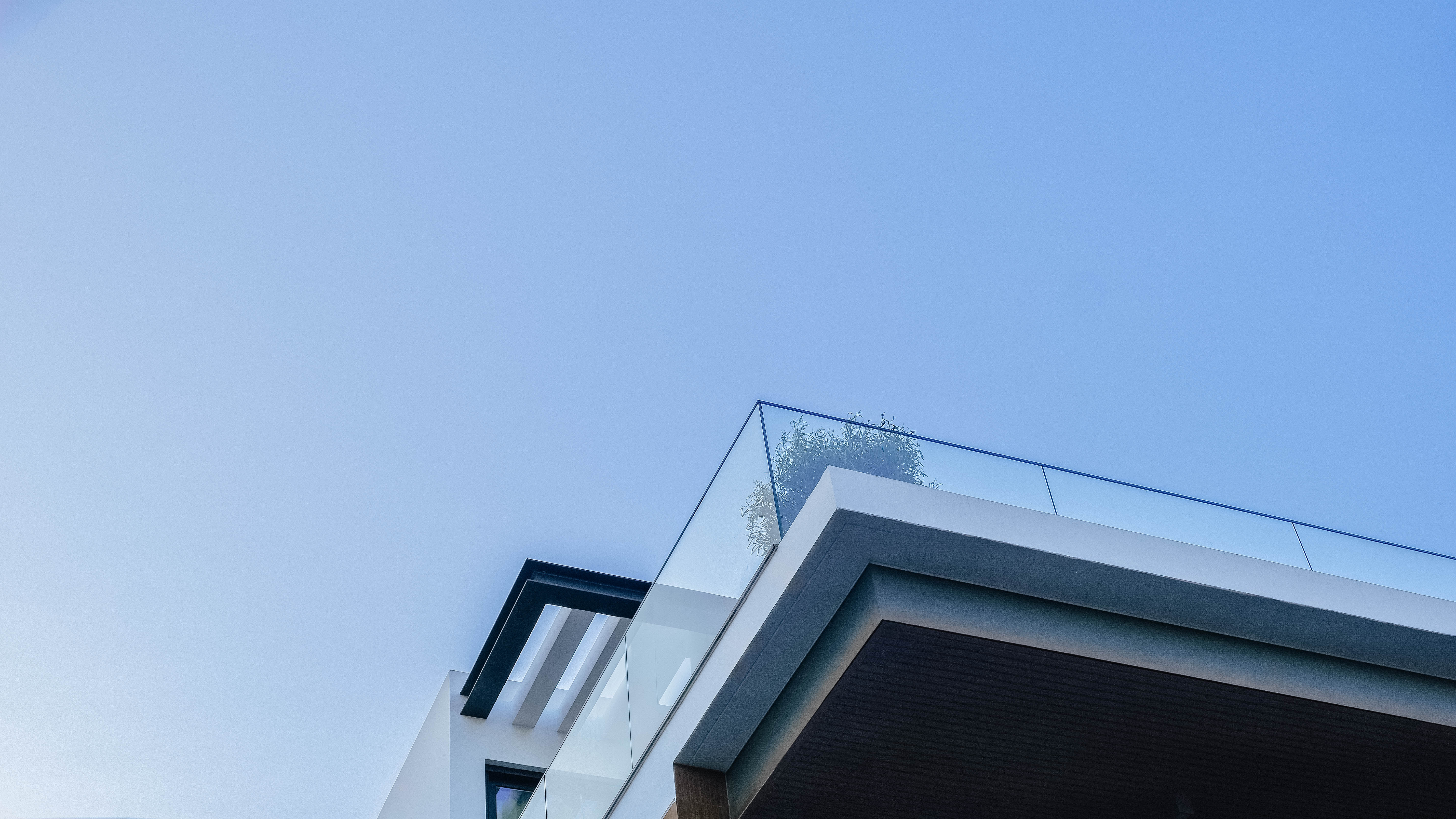 Detalhe de um canto de varanda de uma casa moderna, com parapeito de vidro, um beiral escuro e céu azul claro. A imagem foca na linha limpa do design.
