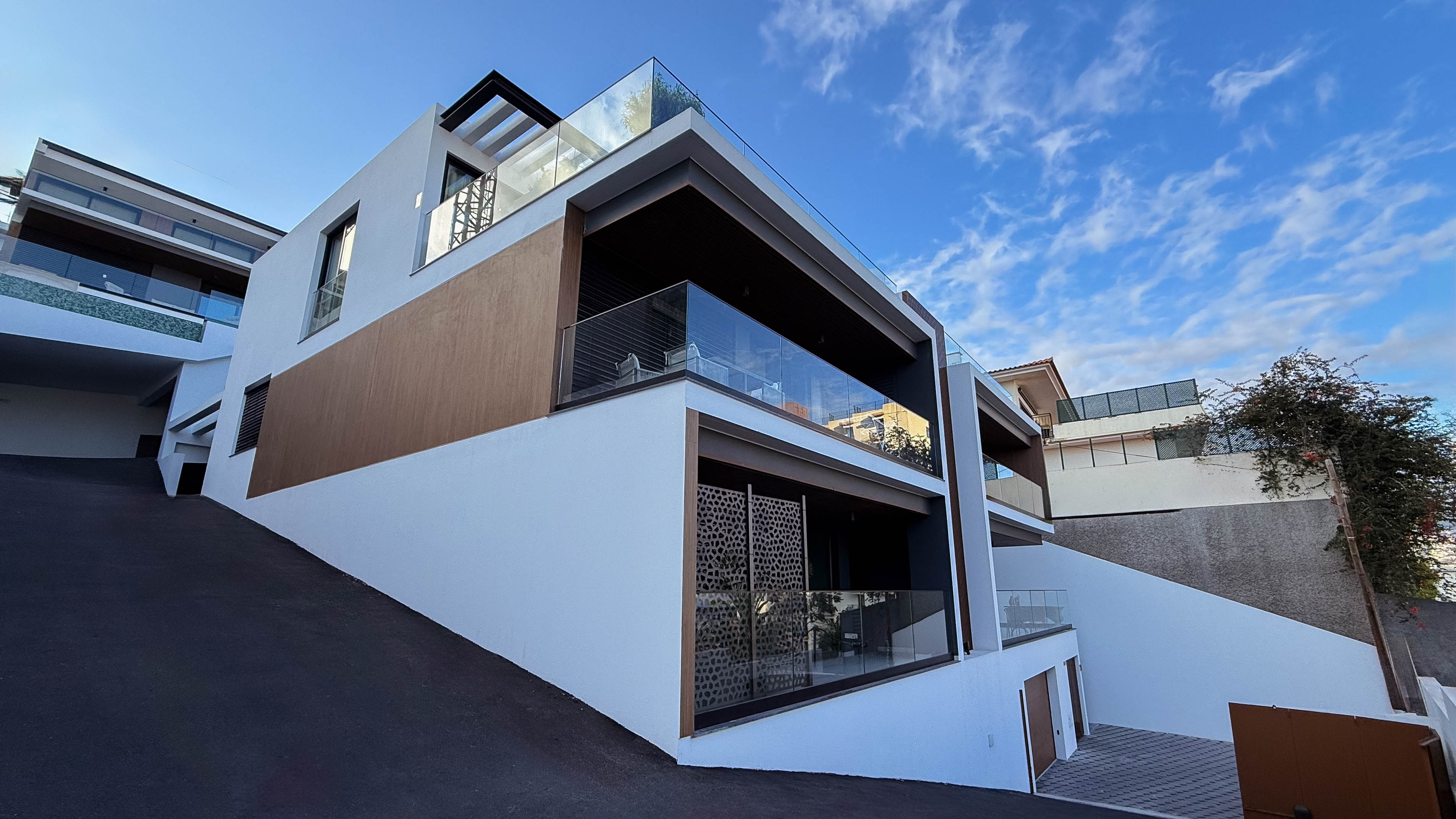Vista exterior de uma casa moderna e luxuosa, com fachada branca, detalhes em madeira e grandes varandas de vidro. A casa está situada numa encosta com uma rampa de acesso, sob um céu azul.