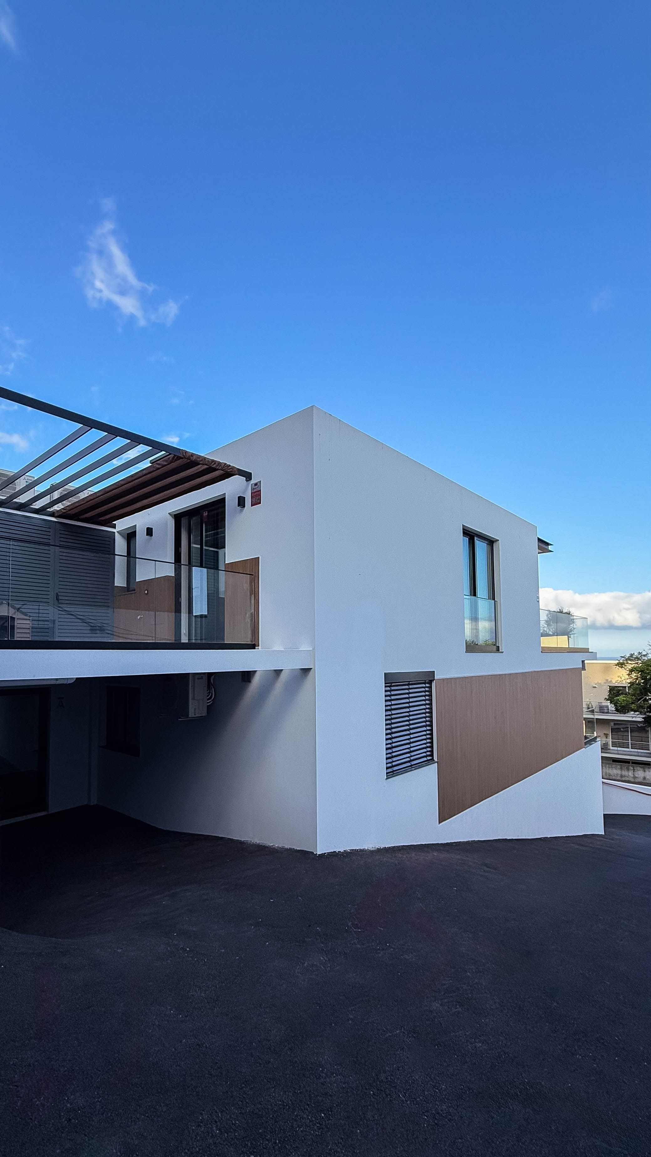 Vista de uma varanda espaçosa de uma casa moderna. A varanda tem parapeitos de vidro, uma pérgola de madeira no topo e a fachada branca da casa, com um pedaço de céu azul visível.