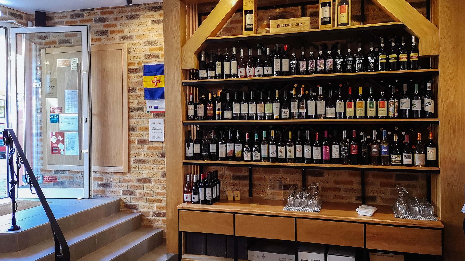 Entrada do Tinta Negra Wine Bar, com a porta aberta, parede de tijolo, degraus em azulejo com um corrimão em ferro com detalhes de folhas de vinha em estilo art déco. Uma prateleira de madeira com variadas garrafas de vinho tinto e alguns copos.