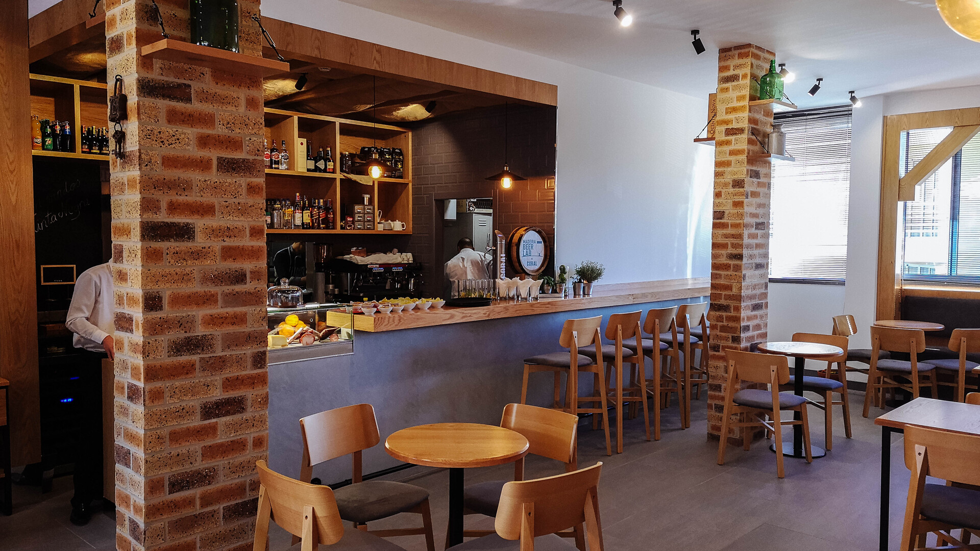 Interior do Tinta Negra Wine Bar com balcão de bar em madeira, prateleiras com bebidas, mesas redondas e cadeiras de madeira, entre colunas de tijolo decorativas.