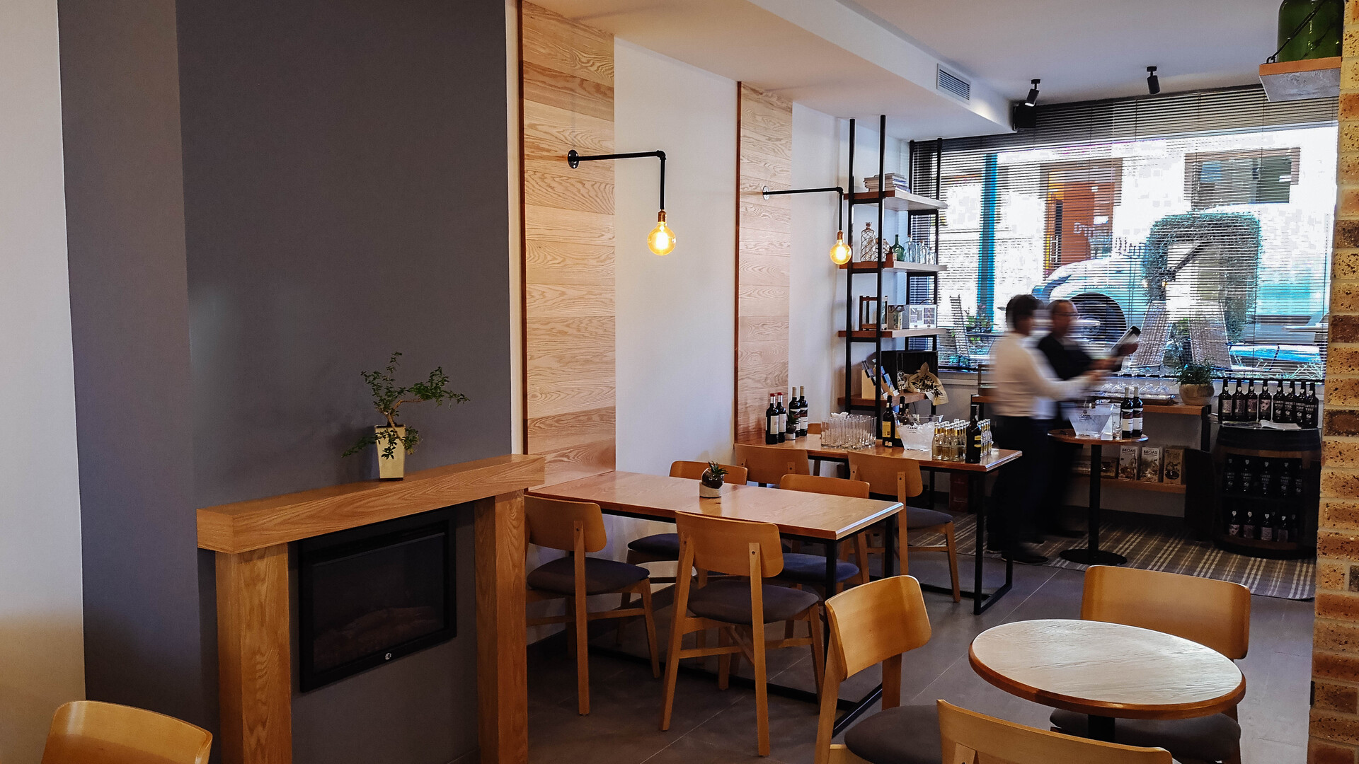 Interior do Tinta Negra Wine bar, com uma coluna cinzenta com uma lareira, na parede branca, umas faixas de madeira com candeeiros de parede iluminam mesas. Ao fundo uma janela e duas pessoas analisam uma garrafa de vinho, enquanto outras se estão expostas