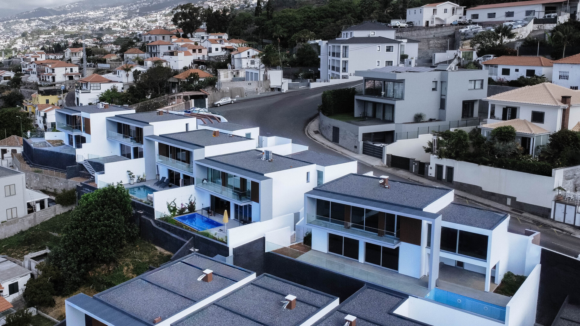 Conjunto de casas de luxo com arquitetura contemporânea e design minimalista, construídas em socalcos. As moradias têm fachadas brancas e cinzentas, grandes janelas de vidro e piscinas privadas, com vista para uma área residencial montanhosa.