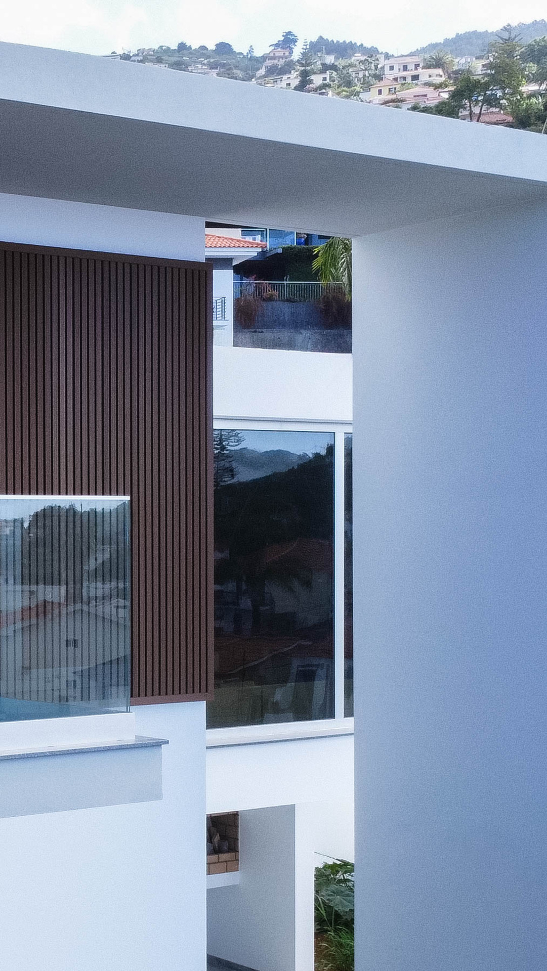 Detalhe da varanda de uma casa moderna, mostrando um pilar de cor clara, painéis de madeira escuros e parapeito de vidro. A imagem mostra um reflexo da paisagem da colina na janela.