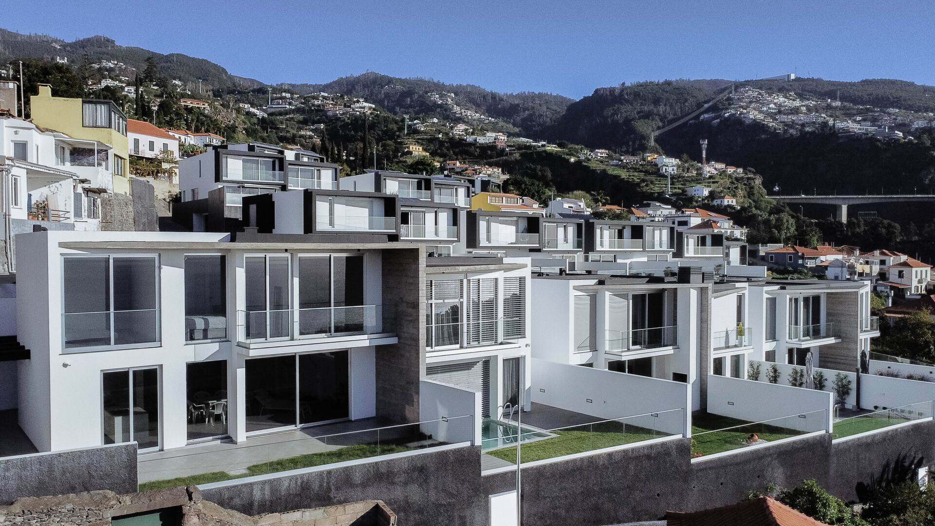 Vista de uma linha de casas modernas e brancas em socalcos numa colina, com grandes janelas, varandas de vidro e jardins privados. A paisagem ao fundo inclui uma área residencial e montanhas.