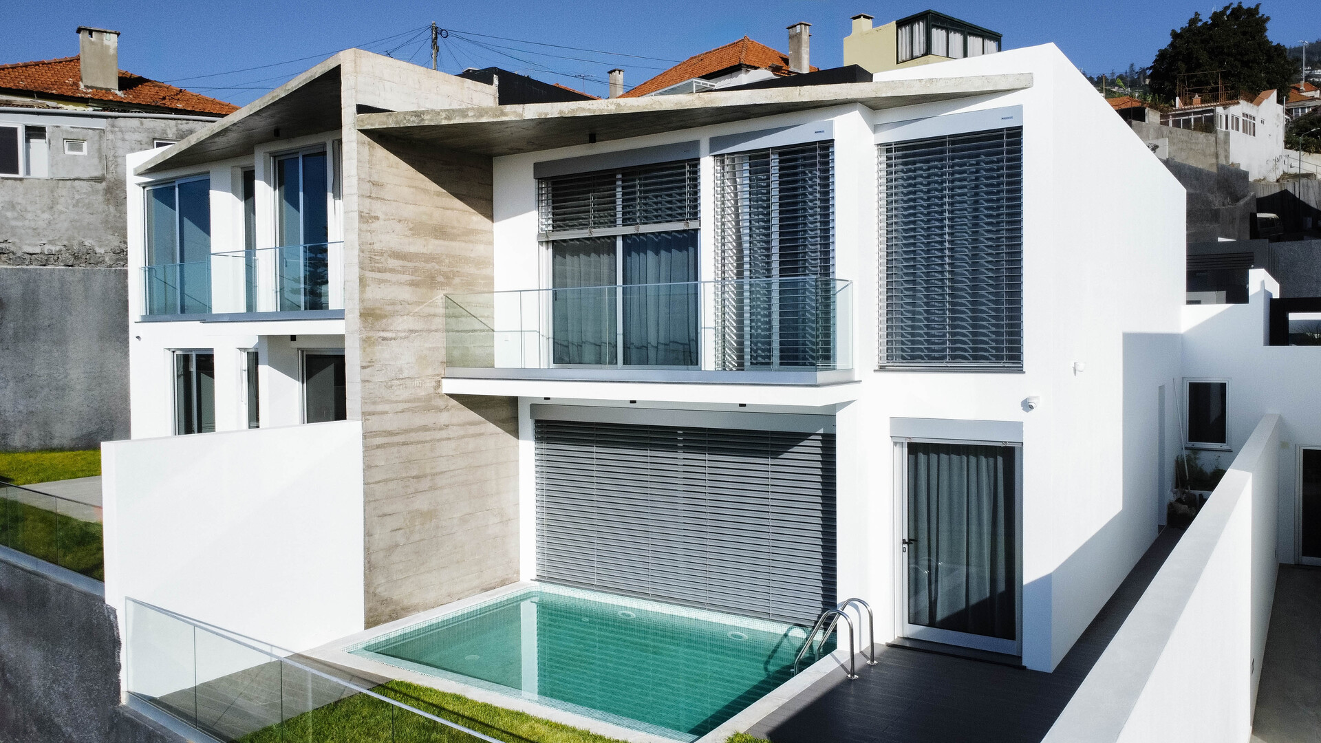 Uma fileira de casas de design moderno, com fachadas brancas e em socalcos, construídas numa encosta. Cada casa tem grandes janelas e varandas de vidro, com pequenos jardins privados e vistas para o vale montanhoso e para a área urbana ao fundo.