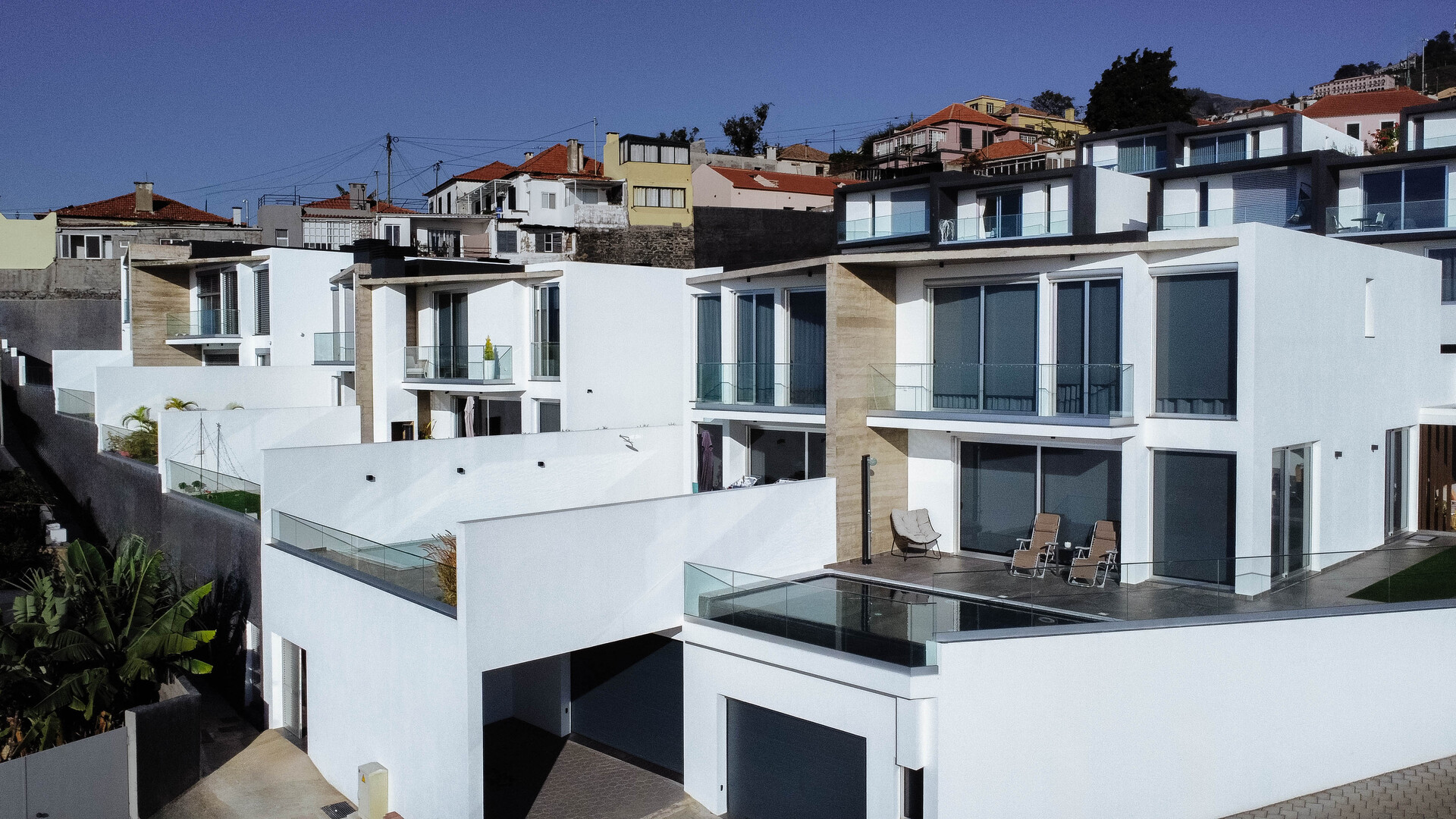 Uma fileira de moradias modernas de dois andares com fachadas brancas e em socalcos, construídas numa encosta com vista para outras habitações. As casas têm grandes janelas, varandas com parapeitos de vidro e garagens integradas ao design.
