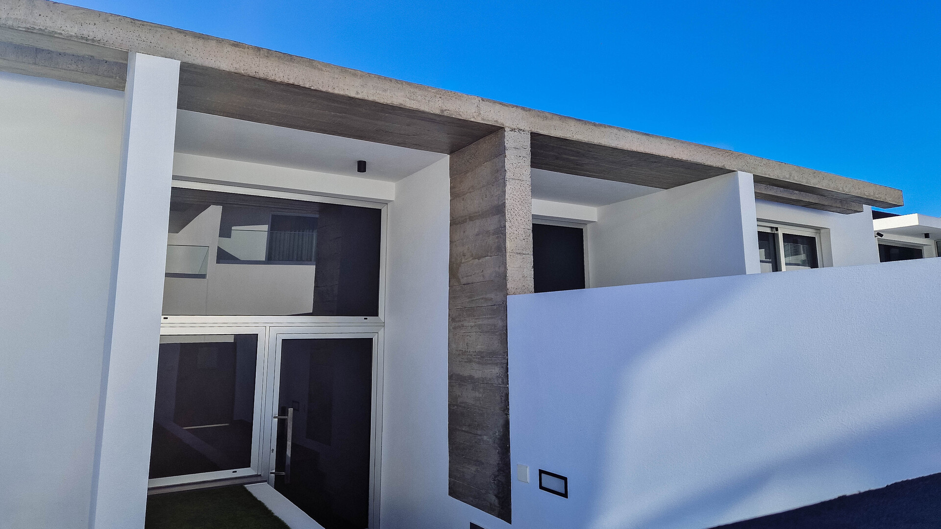 Vista em primeiro plano da entrada de uma casa moderna de cor branca, com uma porta de vidro escuro e uma cobertura de betão saliente apoiada por pilares.