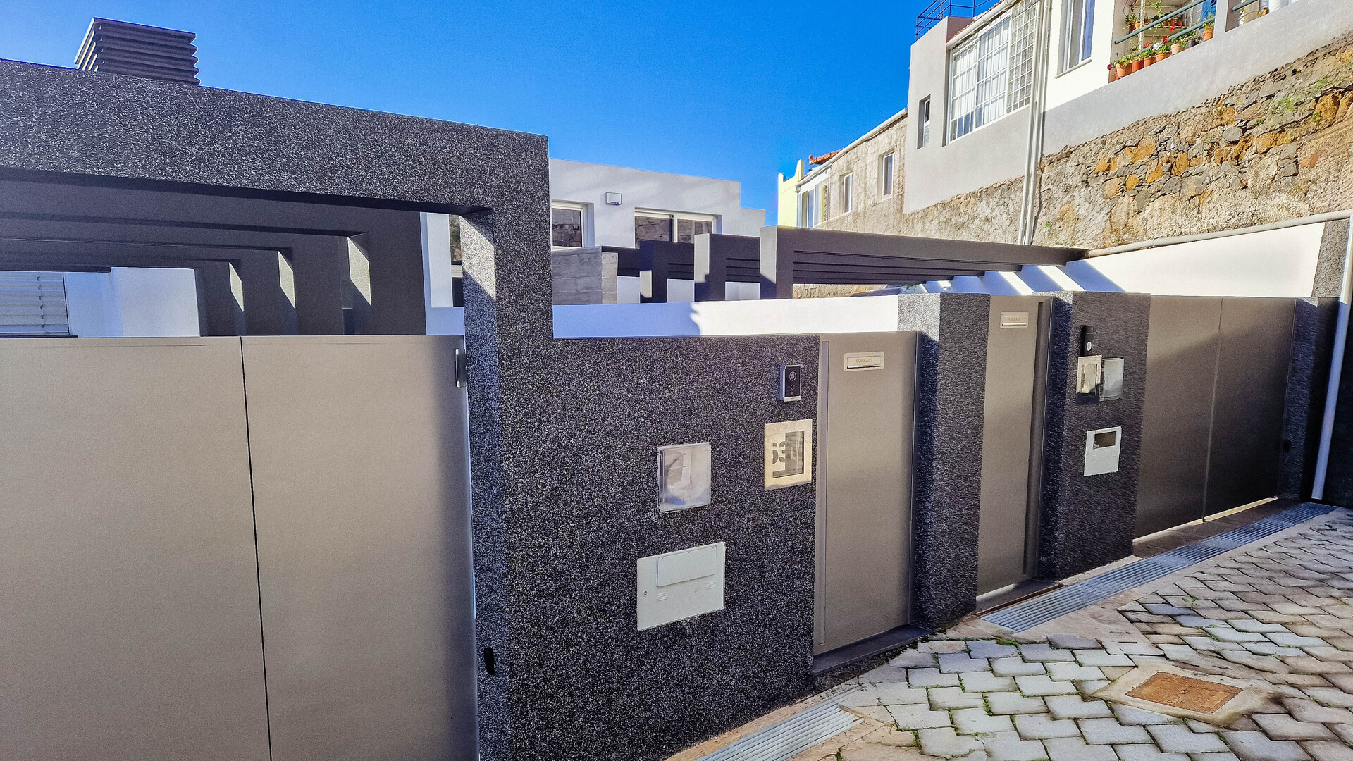 Entrada moderna de uma propriedade, com portões e paredes em cinzento escuro texturizado, contrastando com um céu azul claro. Há um caminho de paralelepípedos e uma parede de pedra no fundo.