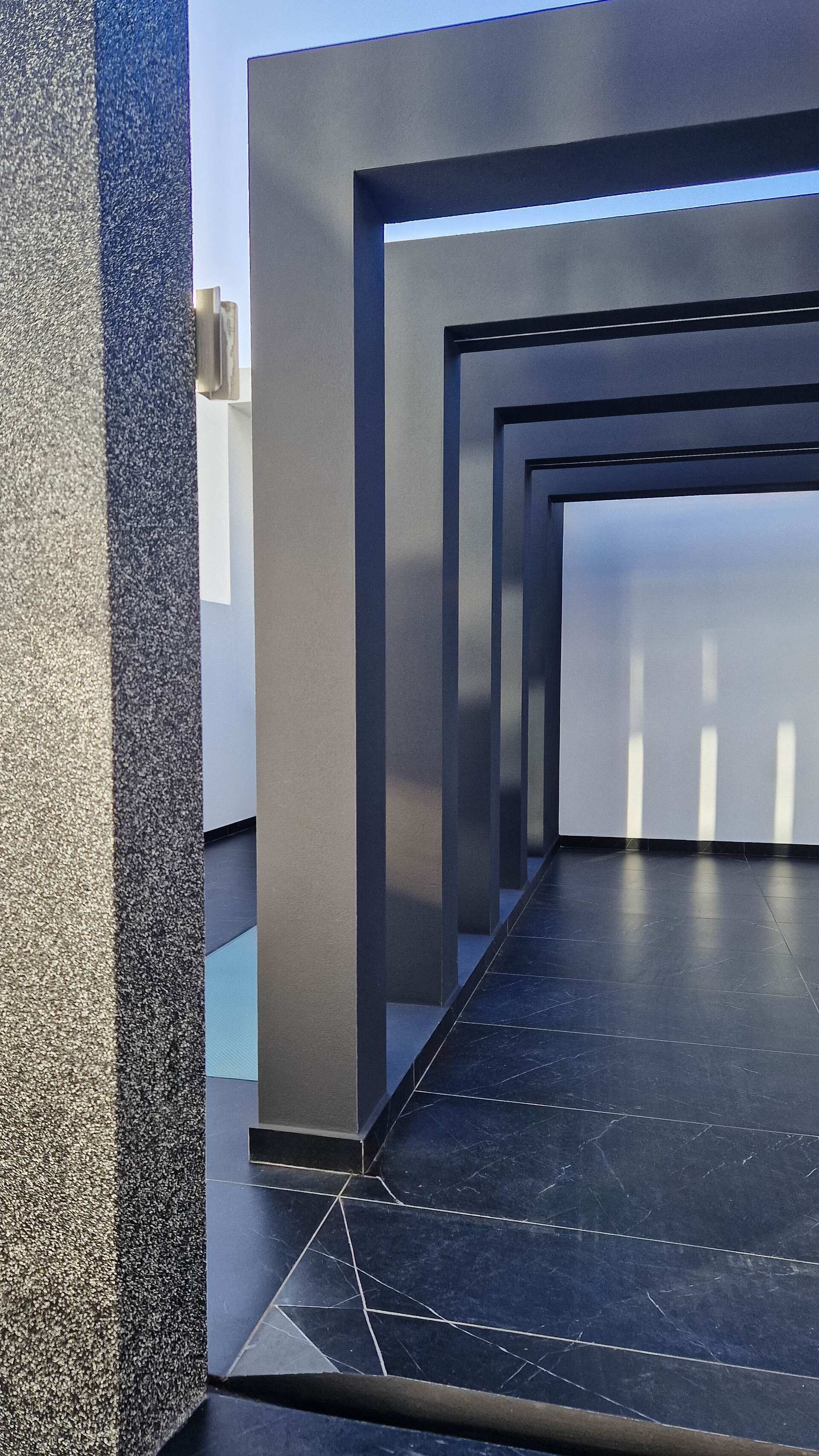 Entrada moderna com uma série de arcos retangulares em tom escuro, criando uma perspetiva profunda. O chão é revestido com mármore escuro. A parede lateral tem uma textura granulada, com luz solar a entrar de cima.