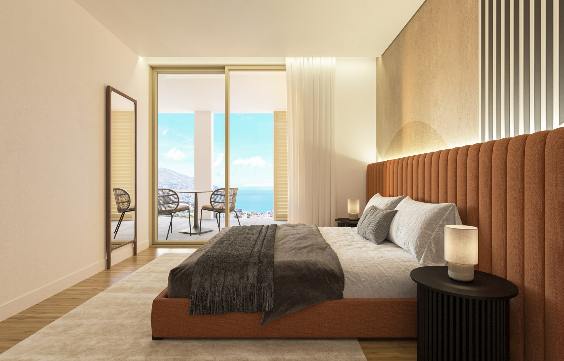 Quarto de dormir moderno e de tons claros e luminoso com uma cama grande de casal com cobertores de tons mais escuros e com um encosto e estrado de tons castanhos. Contém um espelho e acesso envidraçado para um espaço exterior com mesas e vista sobre o mar