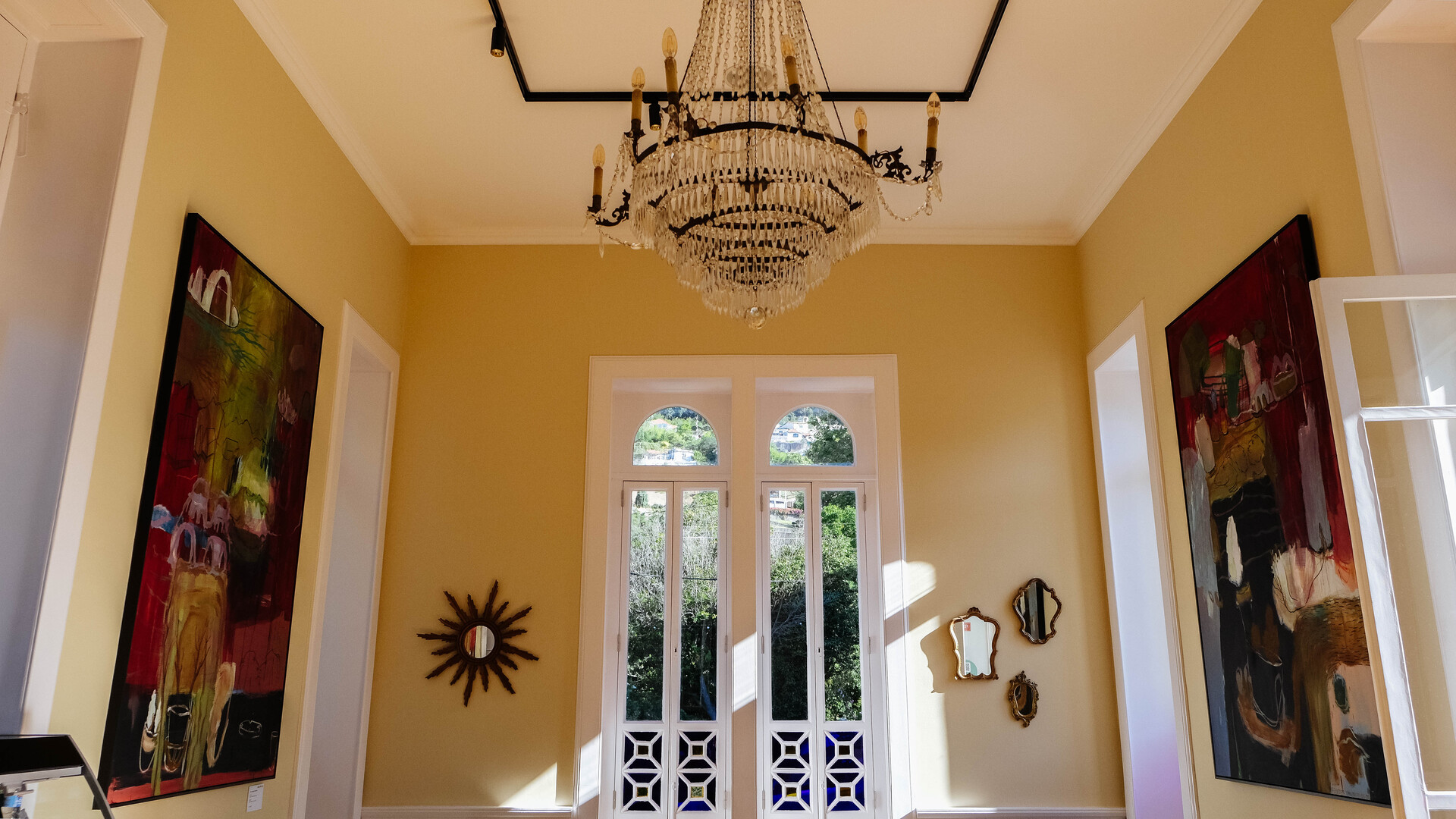Interior com paredes amarelas, teto com lustre de cristal, porta de vidro com detalhes em branco, e obras de arte coloridas penduradas nas paredes.