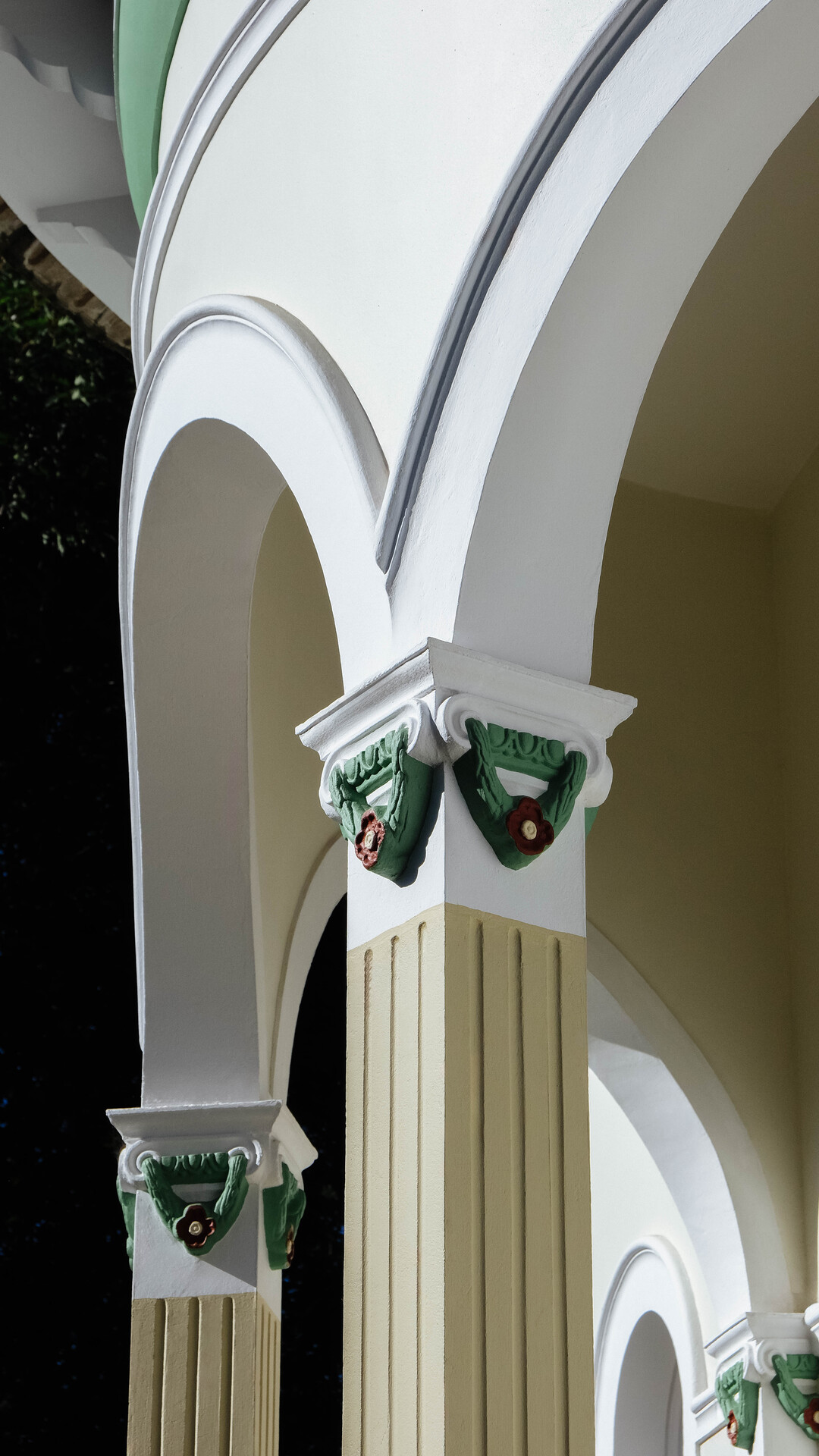 Detalhe de uma coluna decorada com elementos verdes e vermelhos, em uma estrutura com arcos brancos e detalhes clássicos