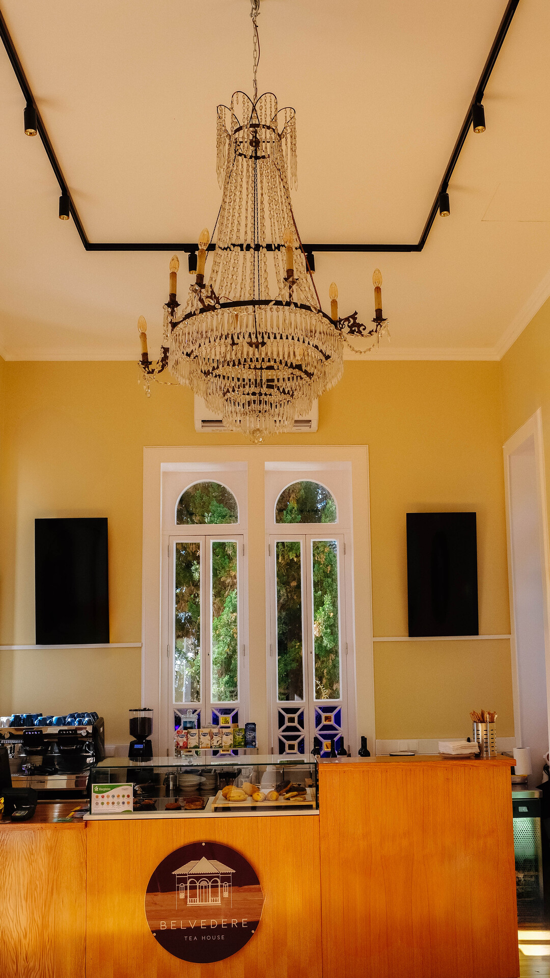 Interior com paredes amarelas, lustre de cristais, janelas altas com vista para árvores, e uma bancada com utensílios de café