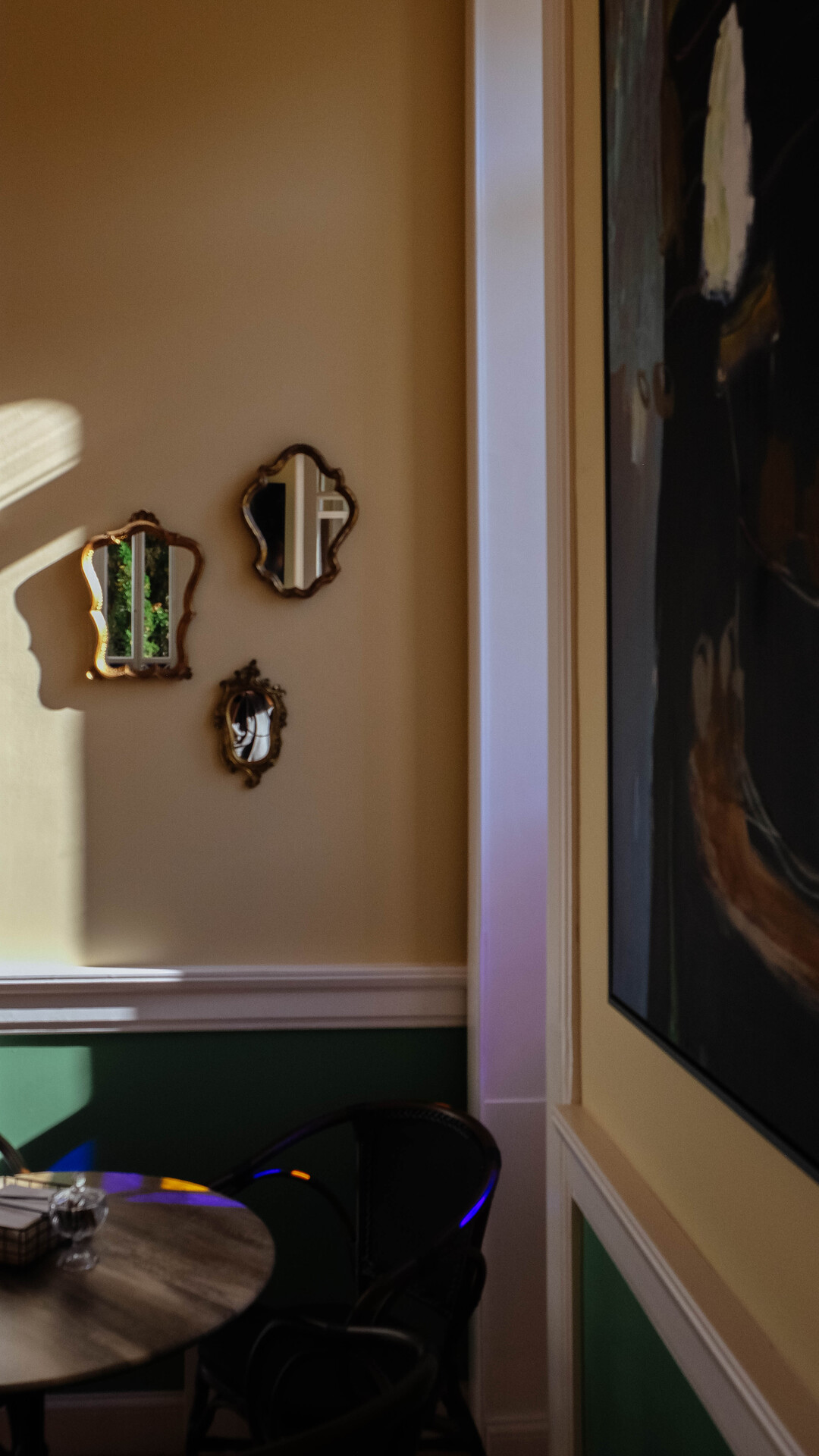 Interior de uma sala com parede bege, decorada com três pequenos espelhos com molduras douradas, uma janela que permite a entrada de luz natural e uma mesa de madeira escura com objetos sobre ela