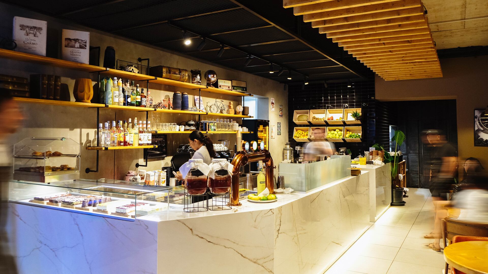 Interior de cafeteria com balcão de mármore branco, expositores de doces, prateleiras de madeira com bebidas e utensílios, iluminação quente e ambiente aconchegante com poucas pessoas