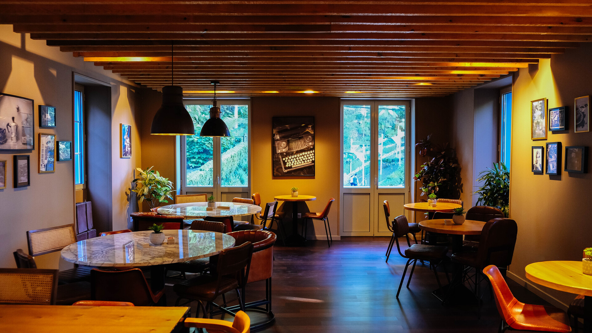 Sala de café ou restaurante com paredes em tom neutro, teto de madeira e iluminação suave, mesas redondas de madeira com cadeiras de couro marrom, decorada com quadros e plantas ao redor