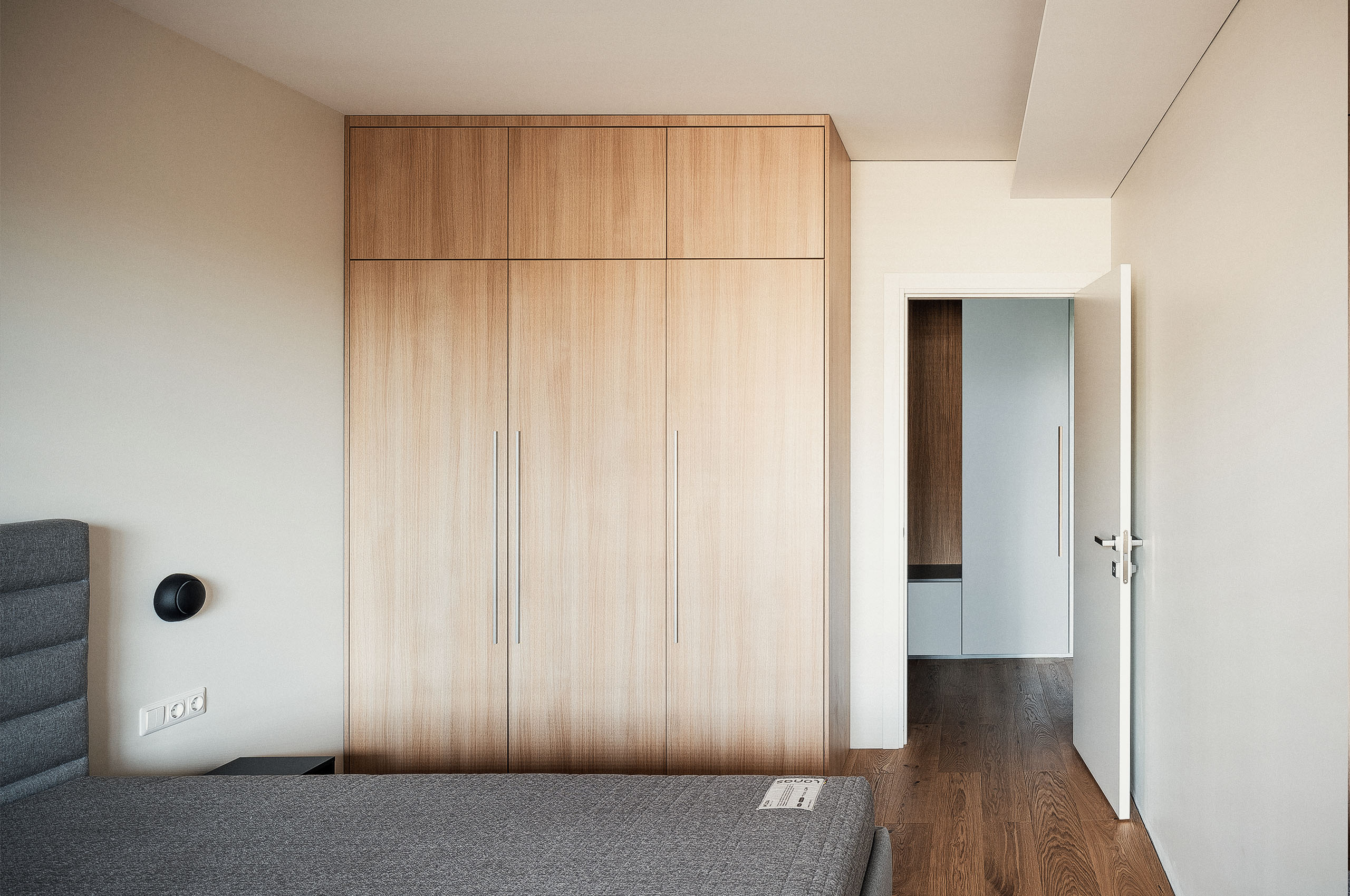 Bright bedroom interior featuring built-in wooden wardrobes, a partially open door, and a grey upholstered bed — designed by TOMA Studio.
