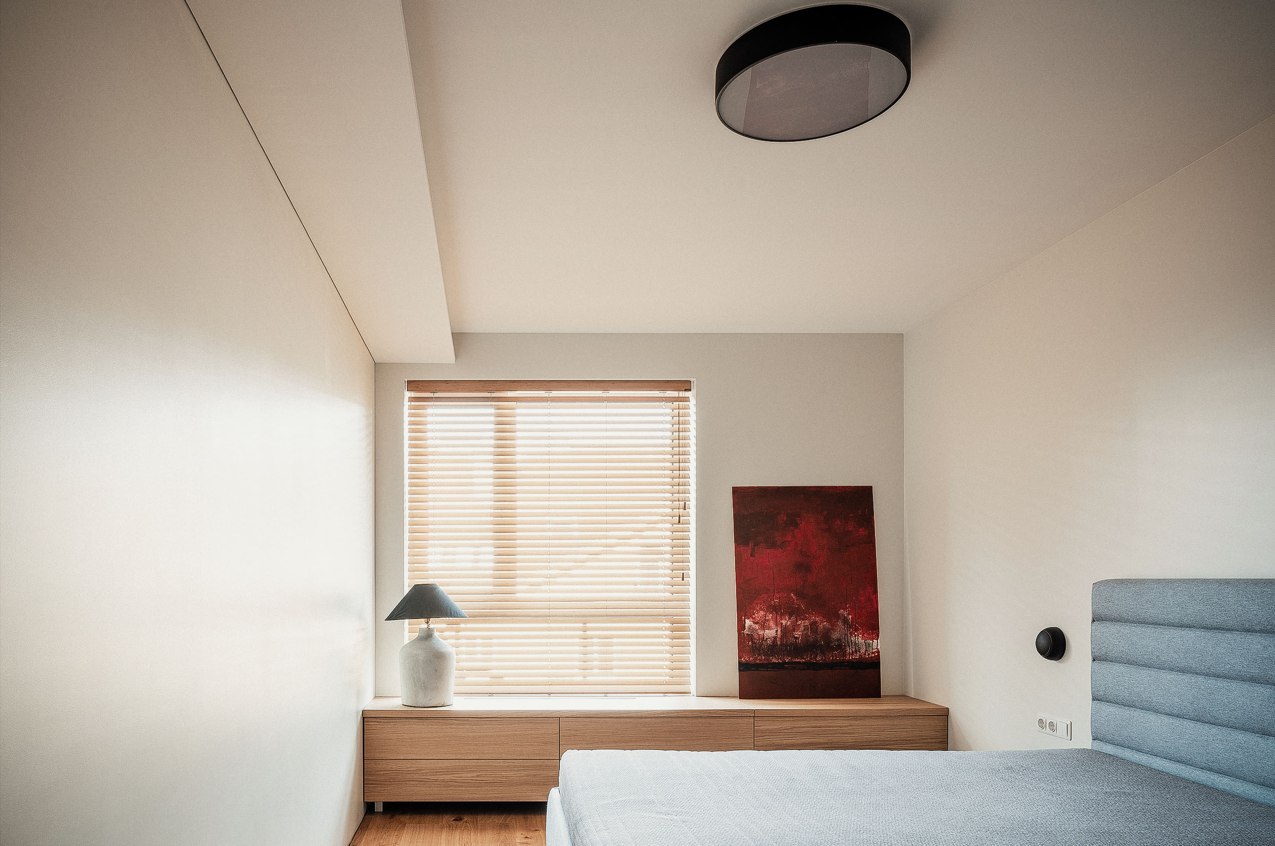 Minimalist bedroom interior with a wooden sideboard, window blinds, a ceramic table lamp, and a red abstract painting next to a grey upholstered bed — designed by TOMA Studio.