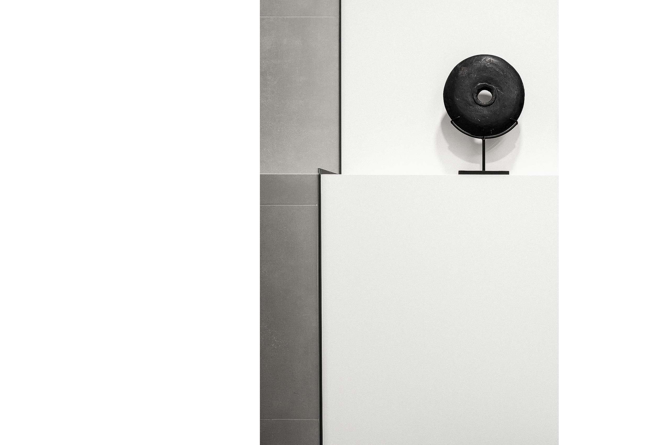 Minimalistische Badezimmerkomposition mit einer skulpturalen schwarzen Steinscheibe auf einer weißen Trennwand.