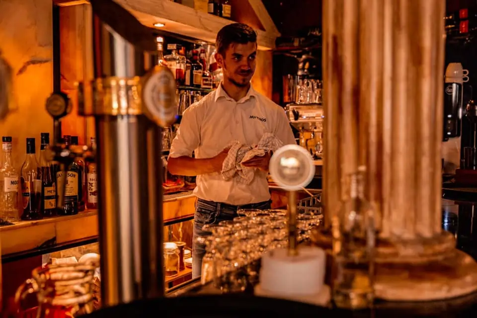 Vitalyc achter de bar van Grieks restaurant Mythos in Hoeven, glazen afdrogend in sfeervolle setting