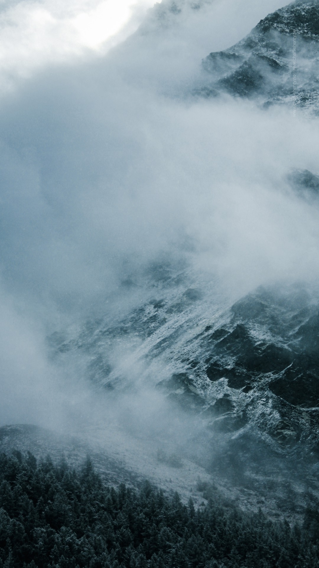 Moody alpine landscape with mist rolling over a forested slope and rugged mountain face in the Altai region — captured by Charlie Henry during a expedition.