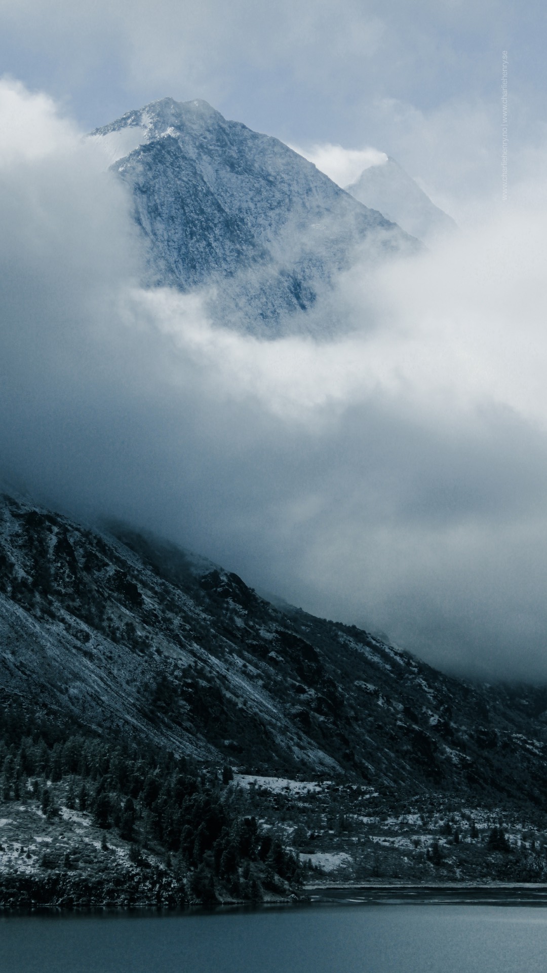 Snow-covered mountain peak emerging through drifting clouds above a dark alpine slope in the Altai Mountains, Russia — photographed by Charlie Henry.
