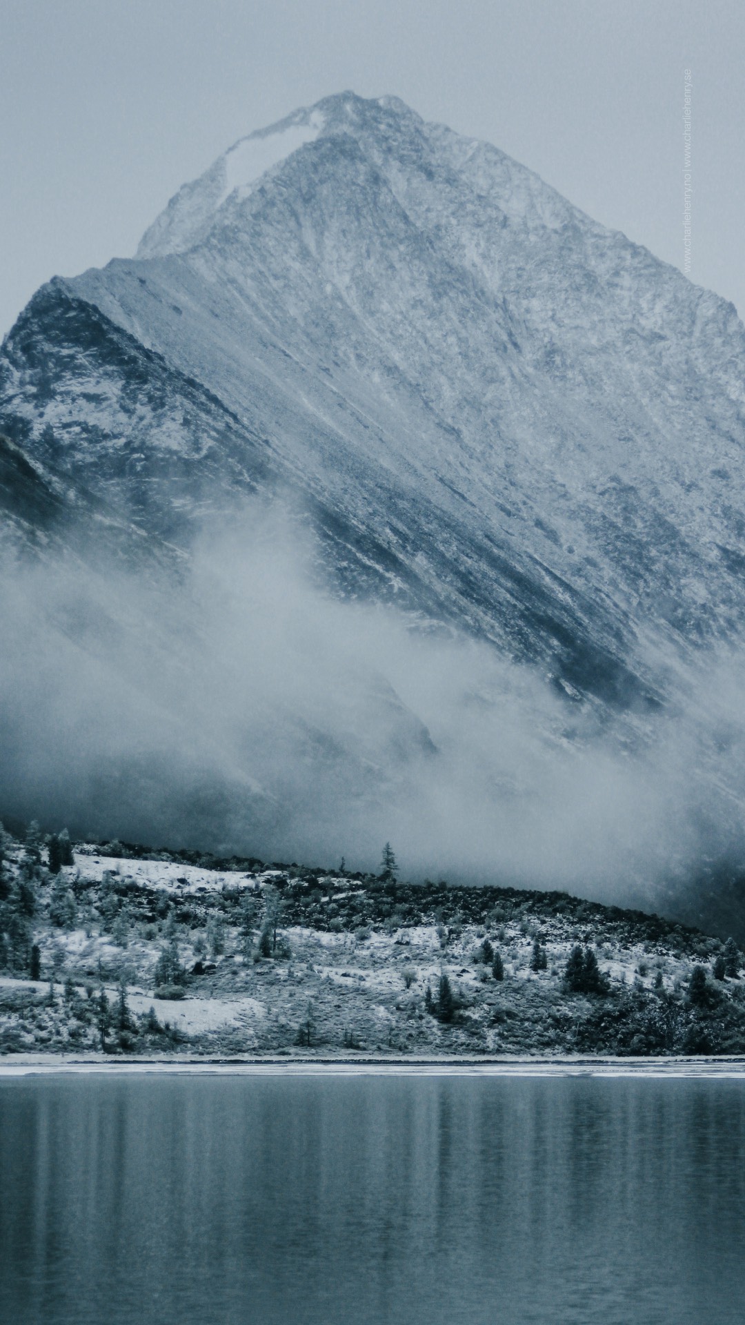 Moody alpine landscape with mist rolling over a forested slope and rugged mountain face in the Altai region — captured by Charlie Henry during a expedition.