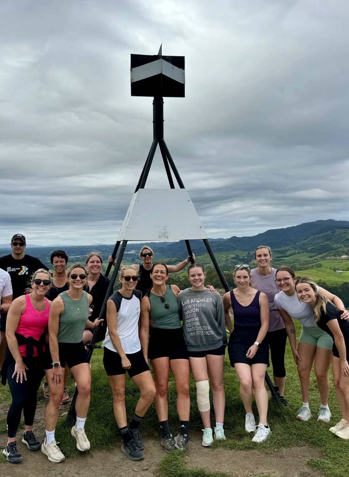 A group of people pose together outdoors near a tall black-and-white triangulation marker on a grassy hill, with rolling green hills and a cloudy sky in the background.