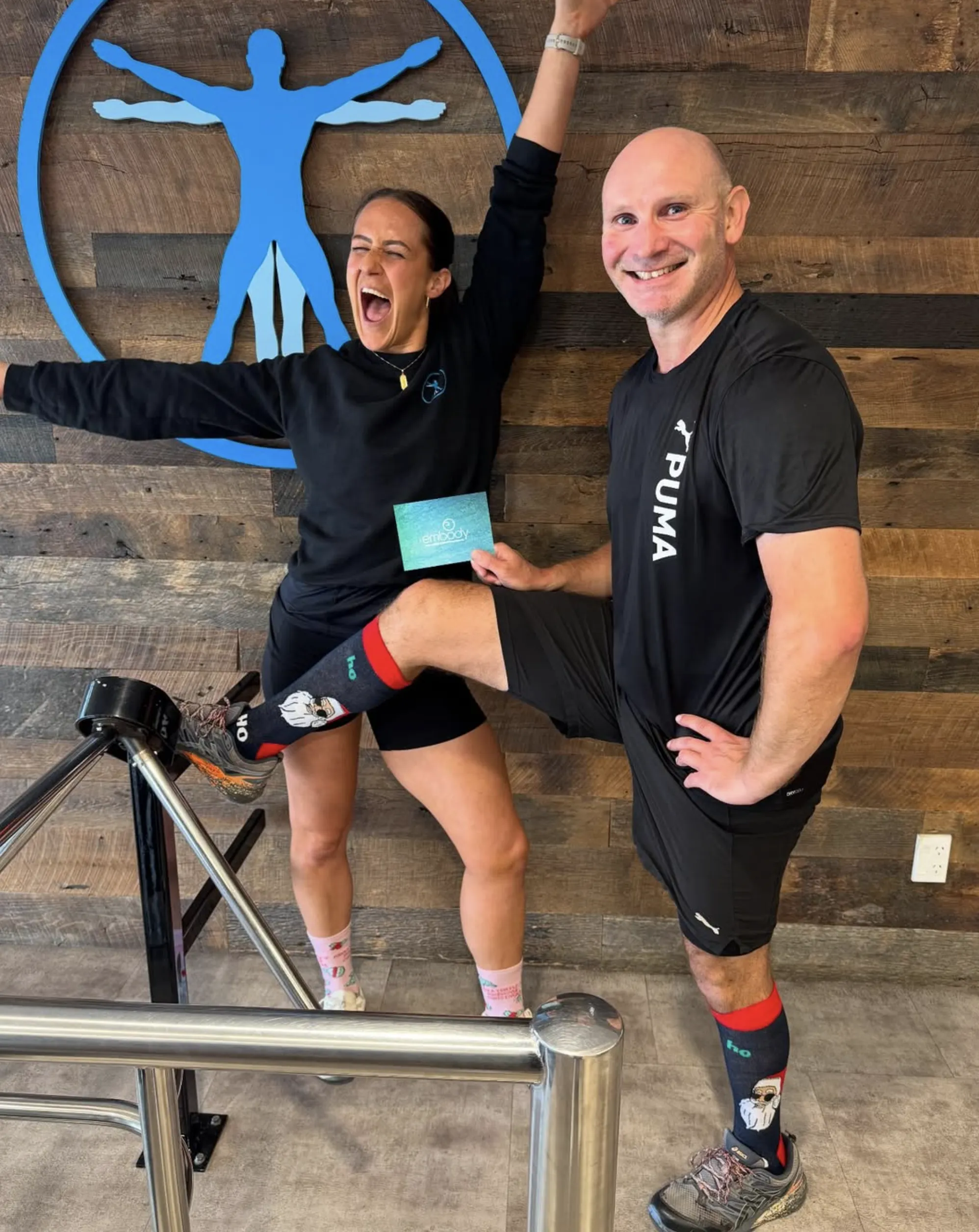 Two people in athletic wear pose playfully indoors; the man smiles with his foot on a railing, while the woman laughs with her arms raised. Both wear colorful socks, standing in front of a blue logo on a wooden wall.
